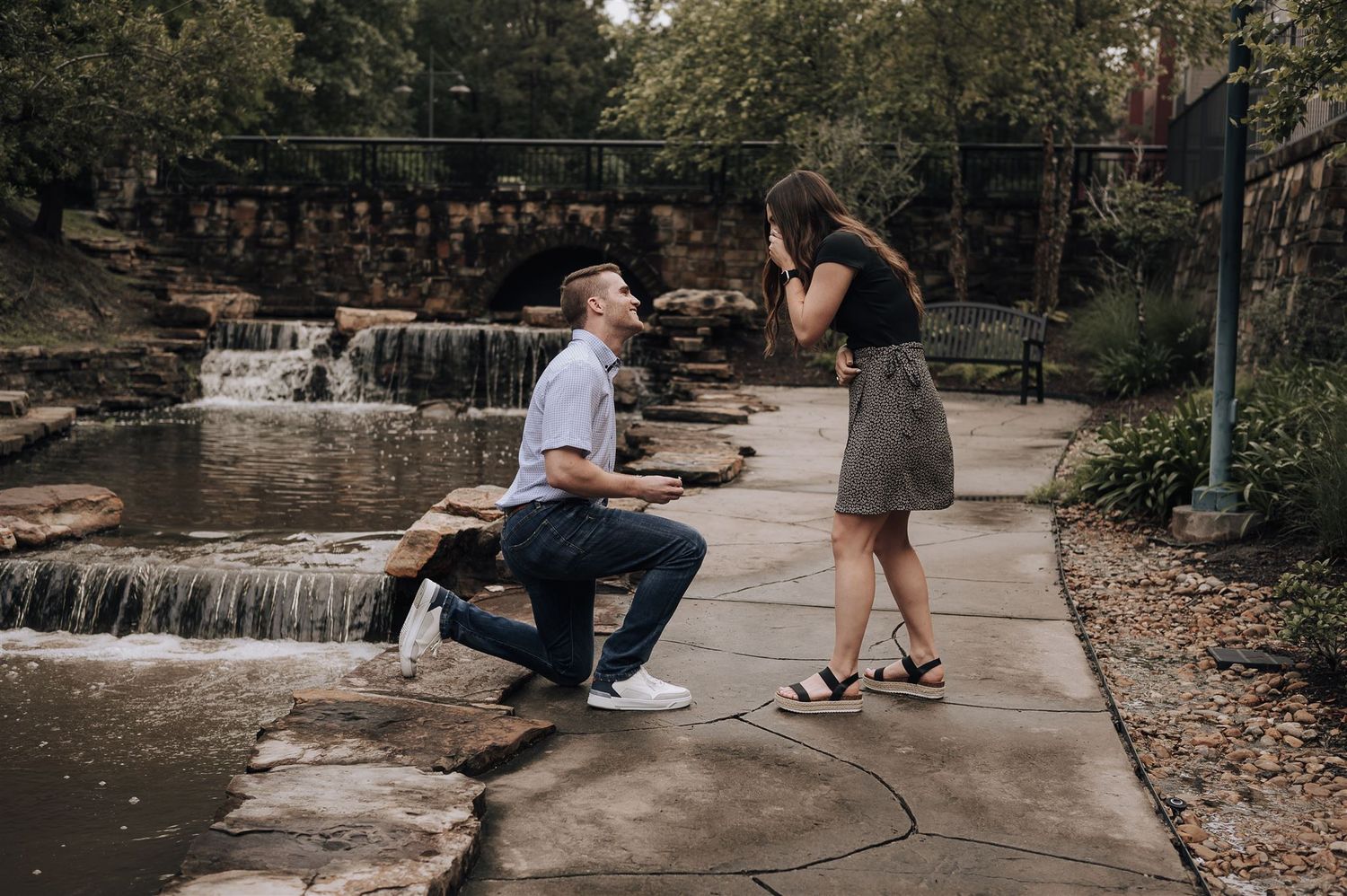 A person proposes near a cascading waterfall on a concrete path surrounded by trees and nature.