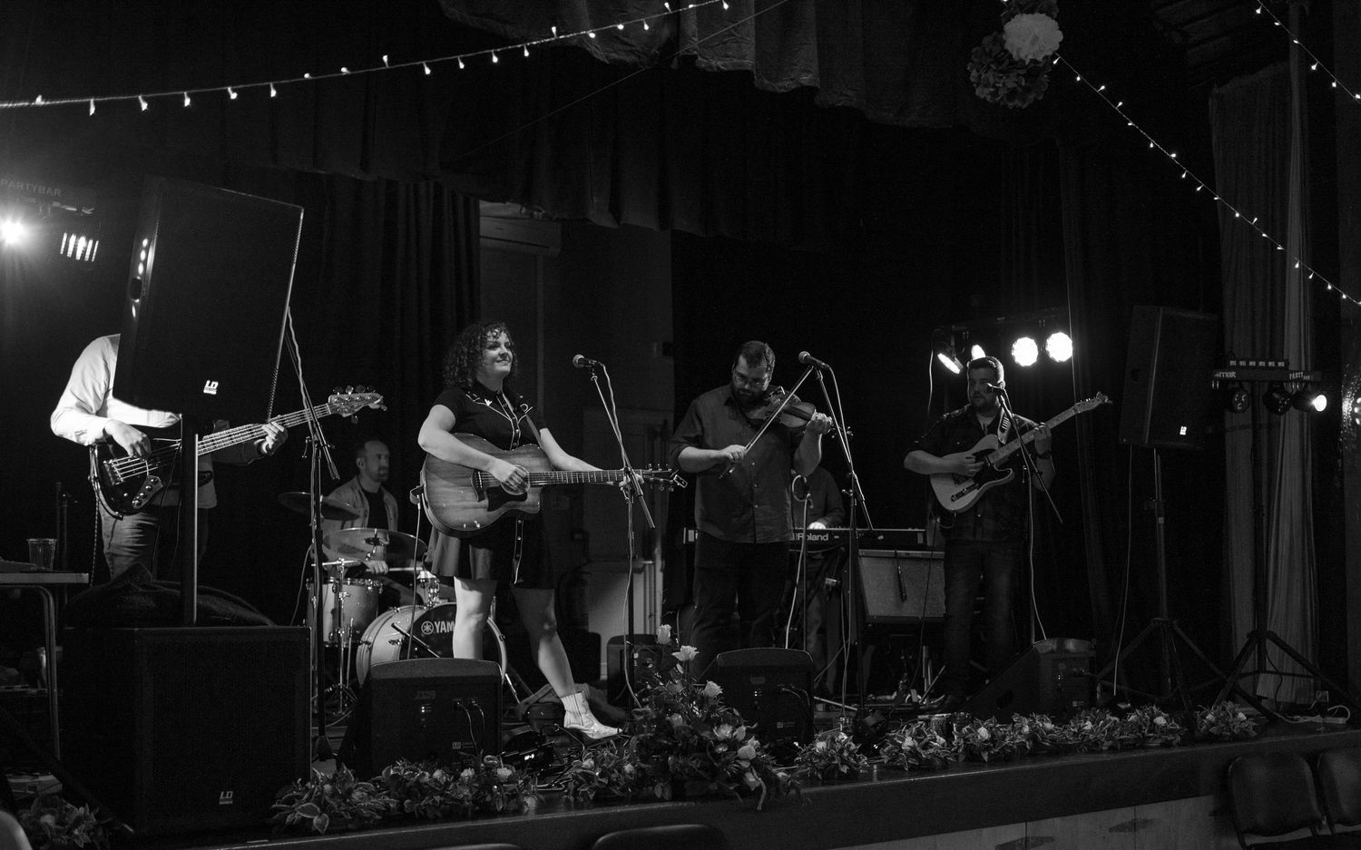Black and white photo of a band performing on stage with string lights overhead.