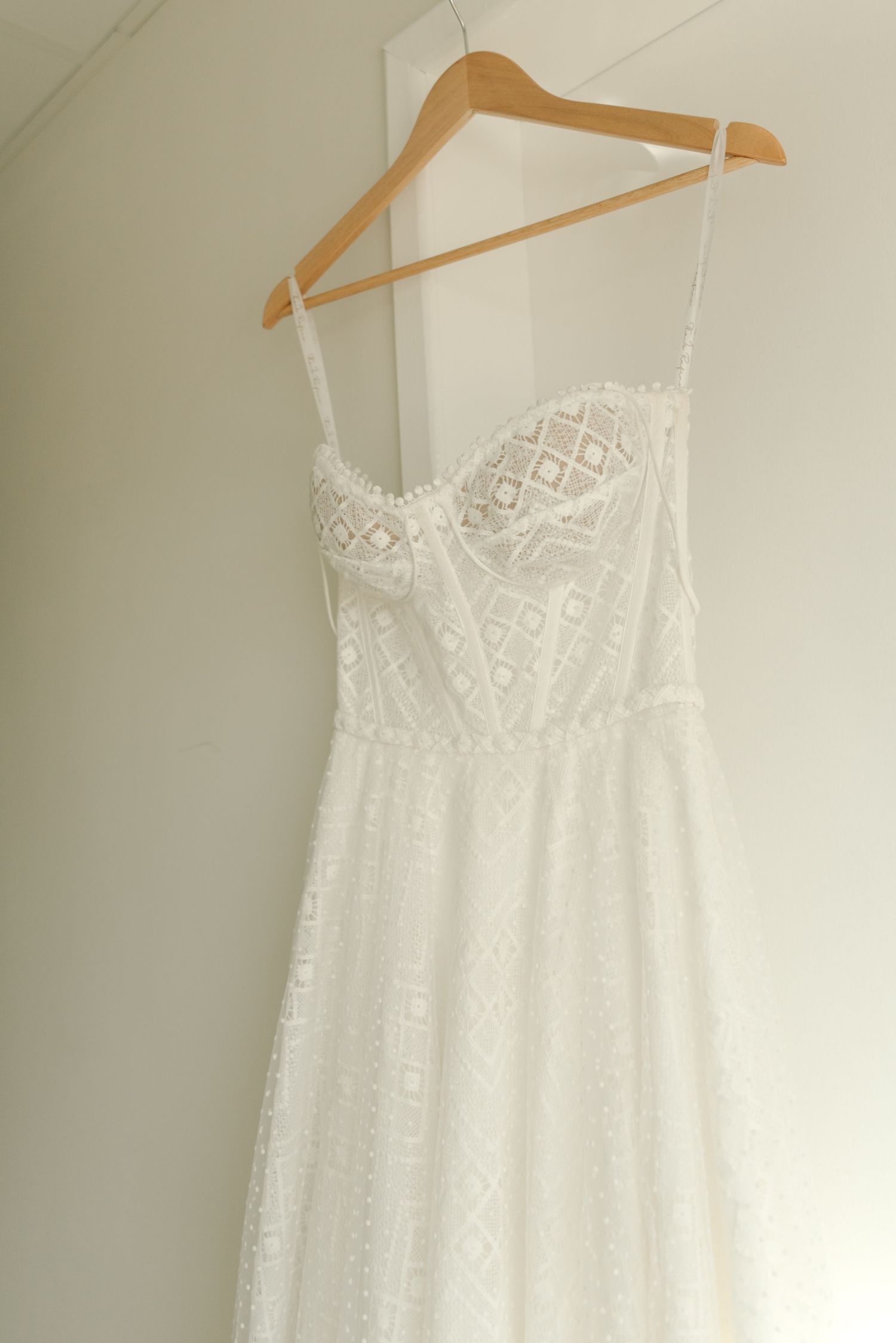 White lace wedding dress with geometric pattern hanging on wooden hanger against light wall.