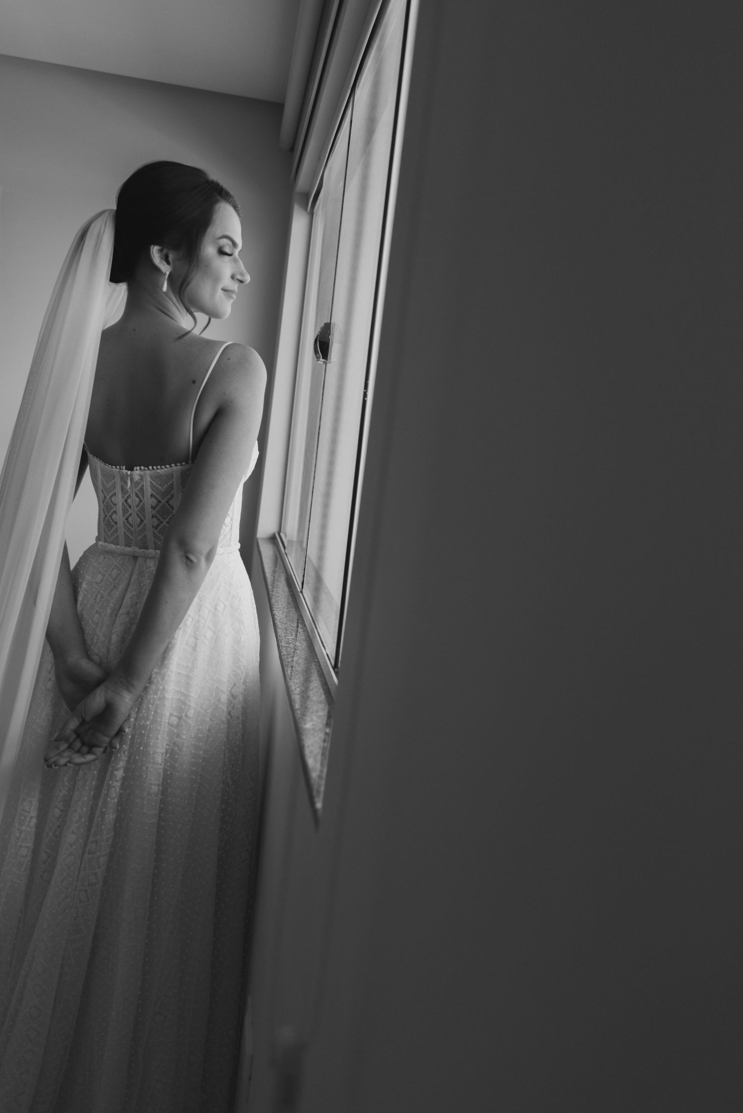 Black and white photo of bride in lace dress looking out window.