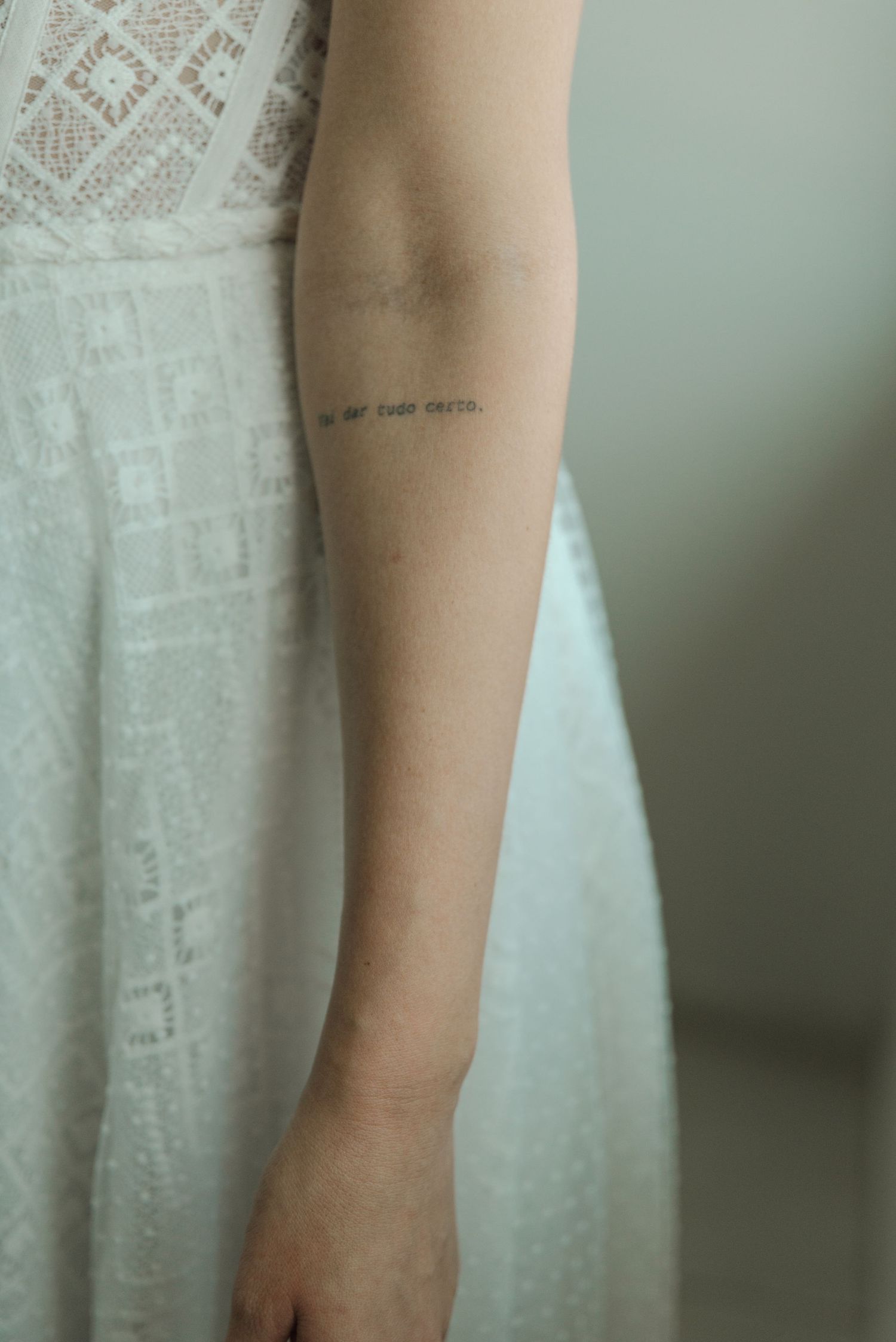 Close-up of small text tattoo on arm with white lace dress detail.