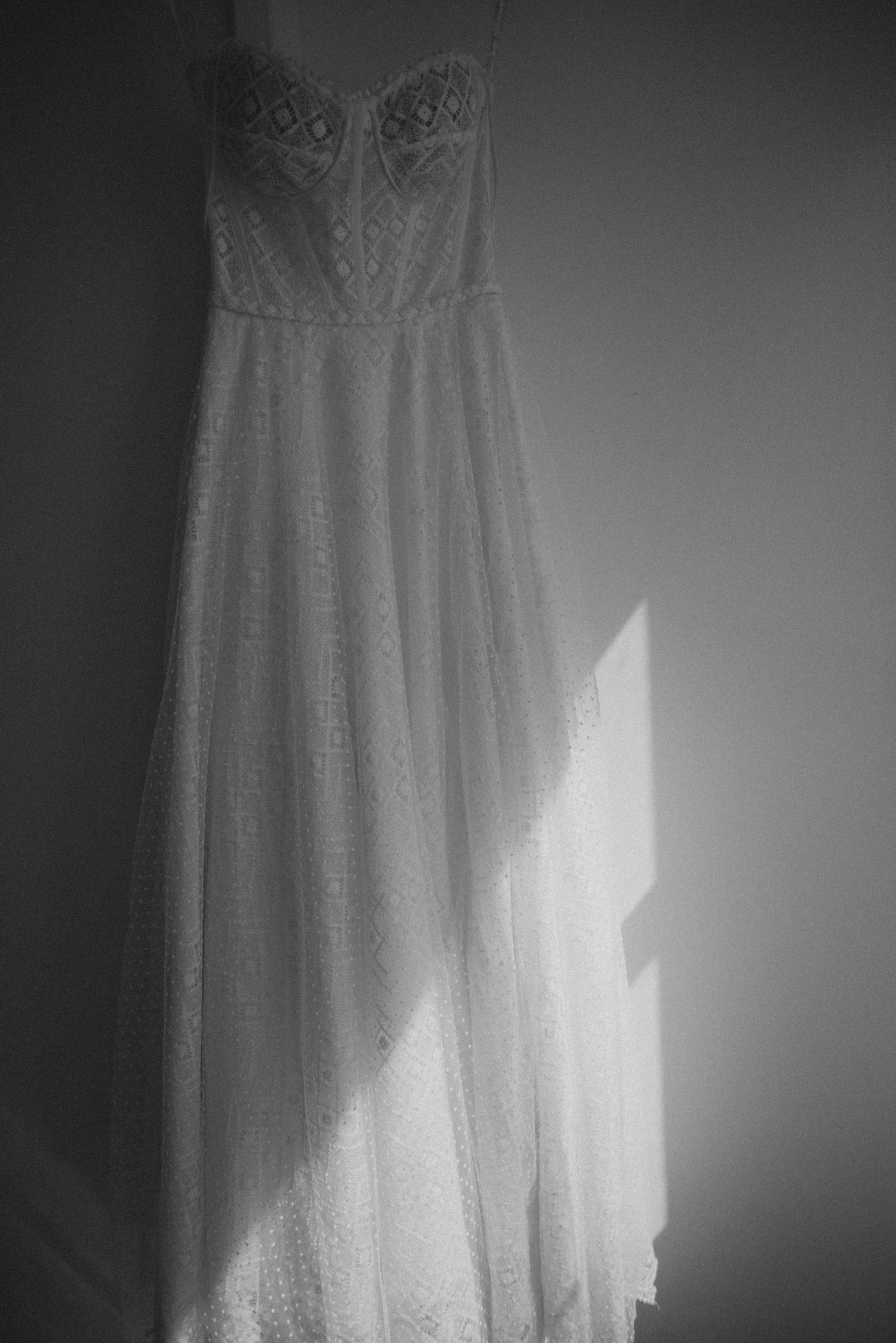 A delicate white lace wedding dress hanging against a wall with dramatic side lighting.