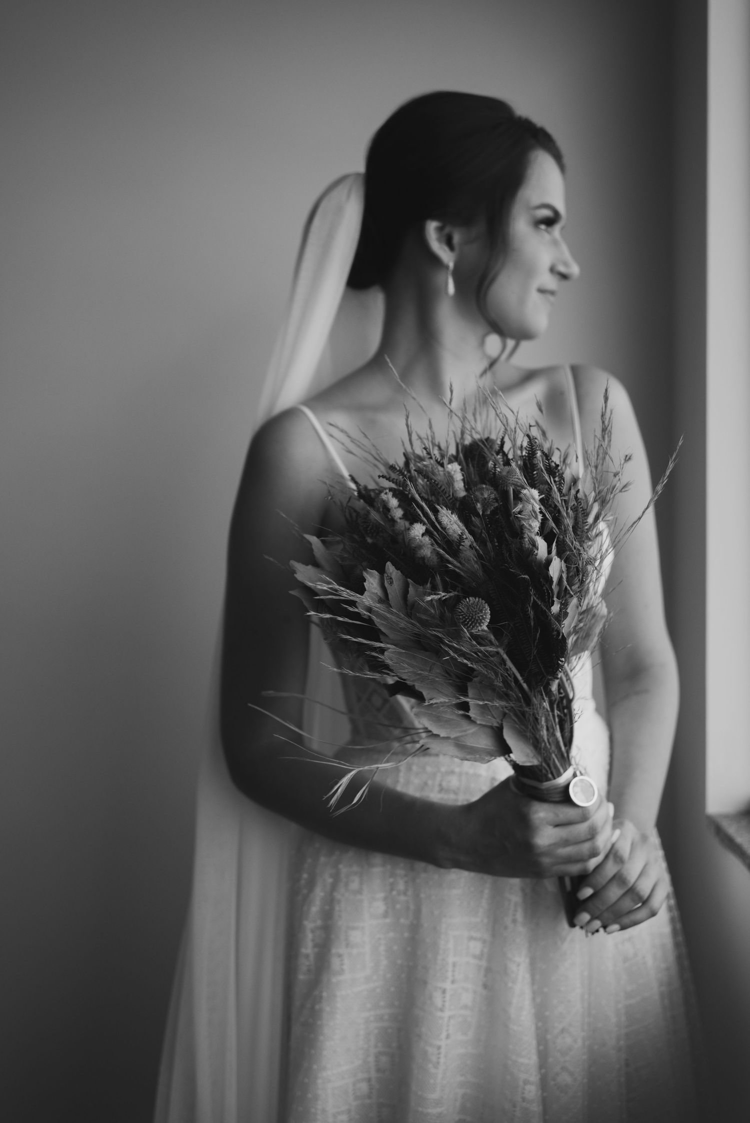 Black and white image of bride holding rustic bouquet near window.