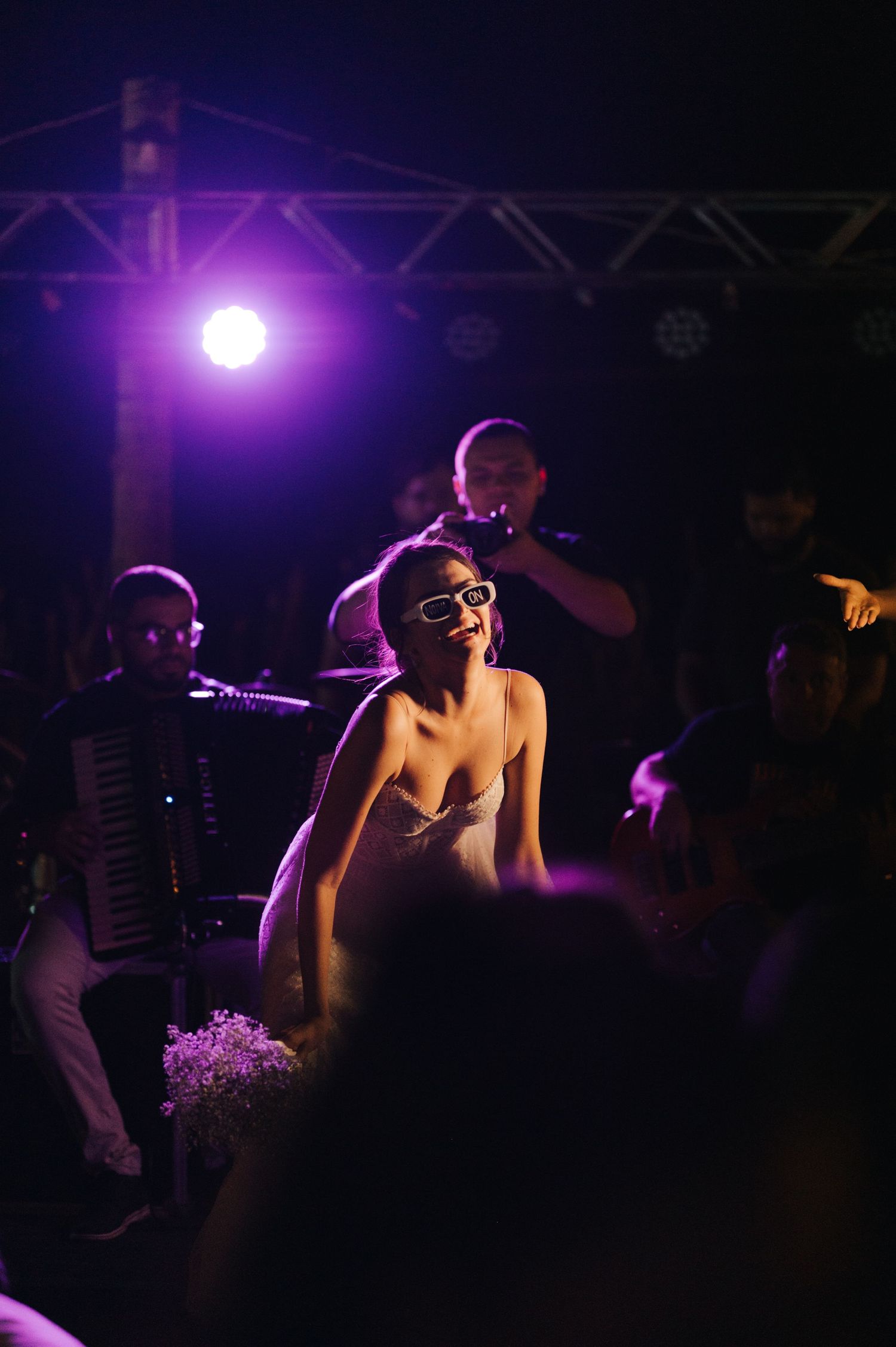 A performer in a white dress illuminated by purple stage lights with musicians in the background.