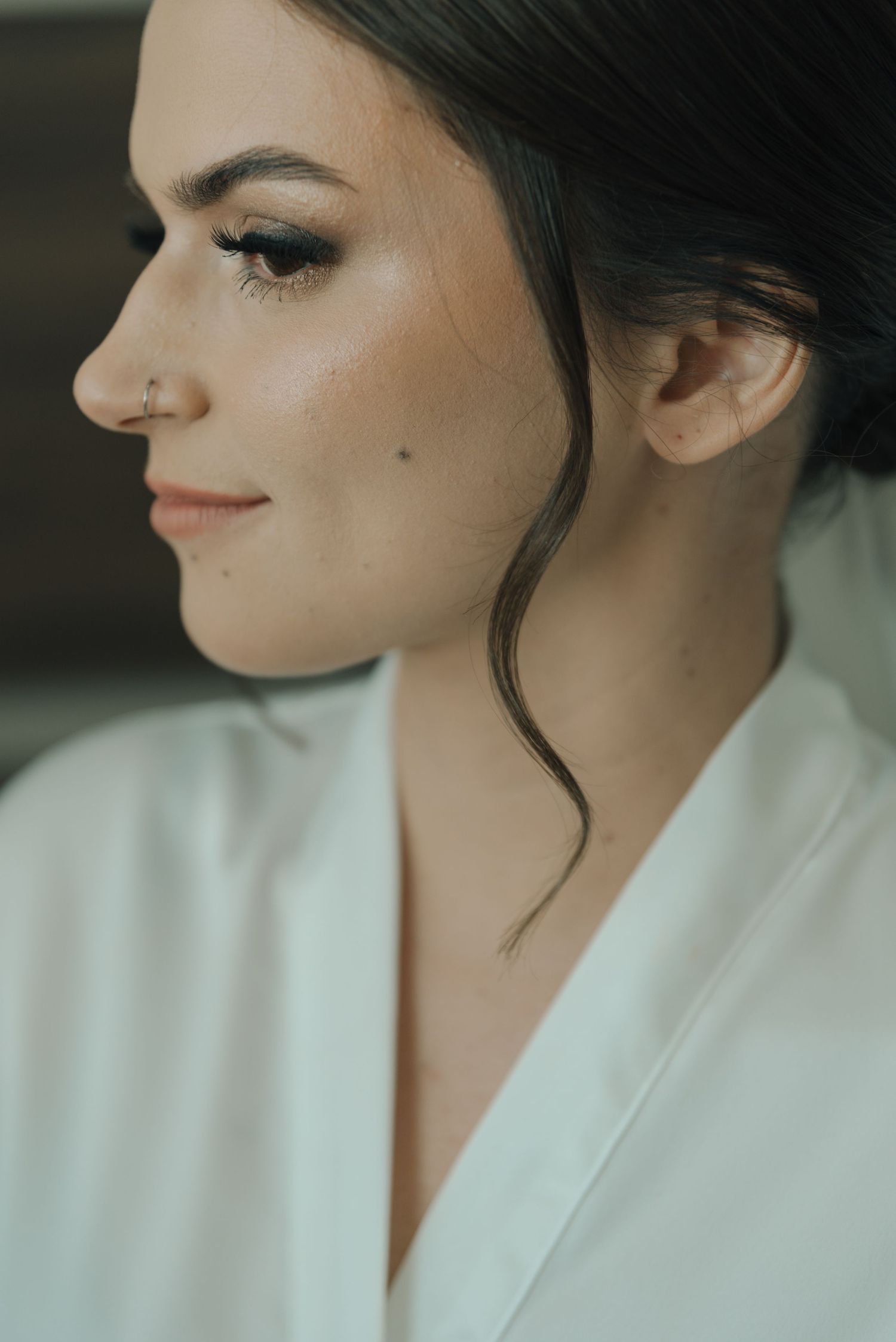 A close-up profile view showing soft makeup and a nose piercing while wearing a white robe.