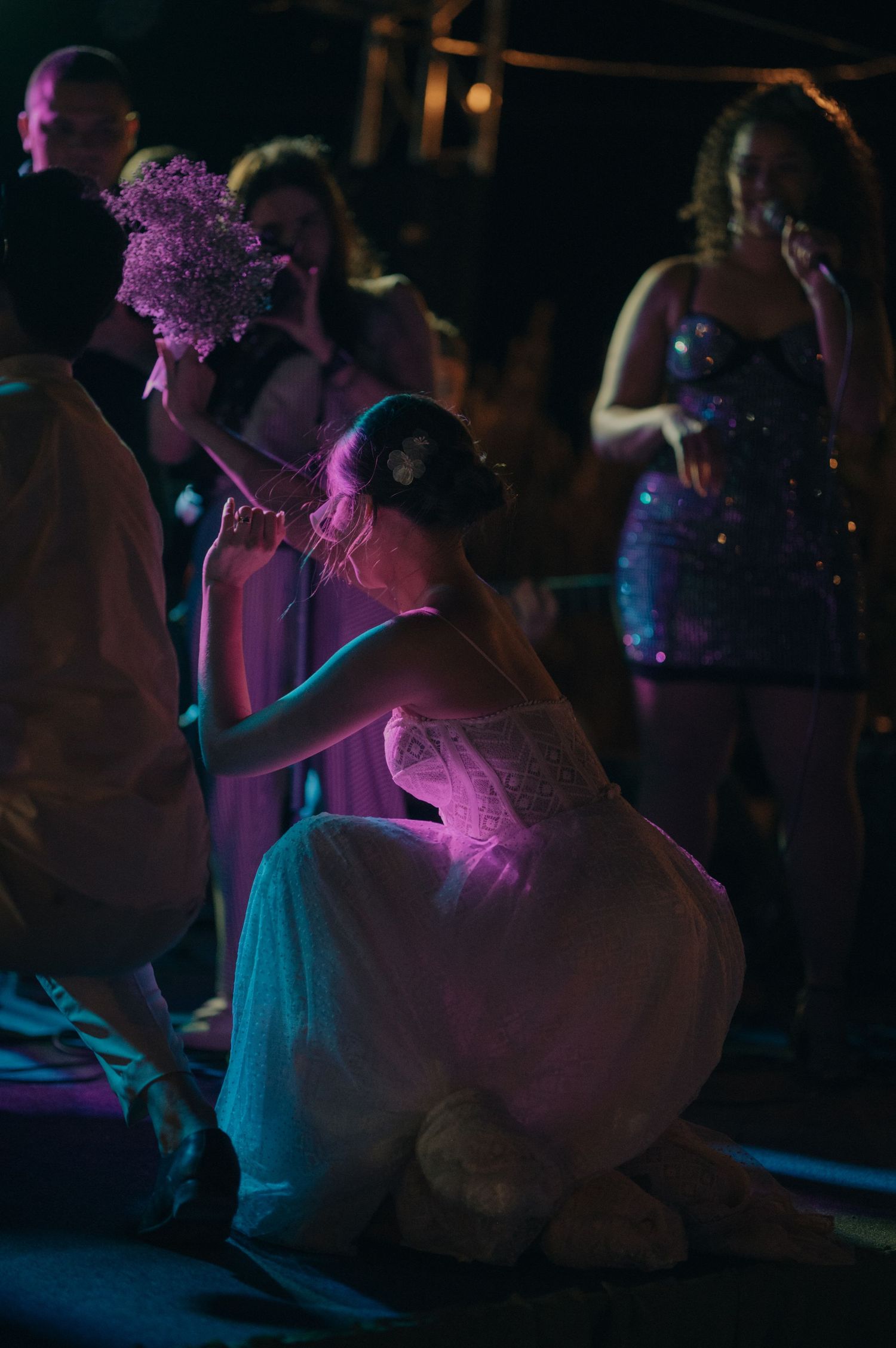 A dancer in a white dress kneels on stage under purple lighting during a performance.