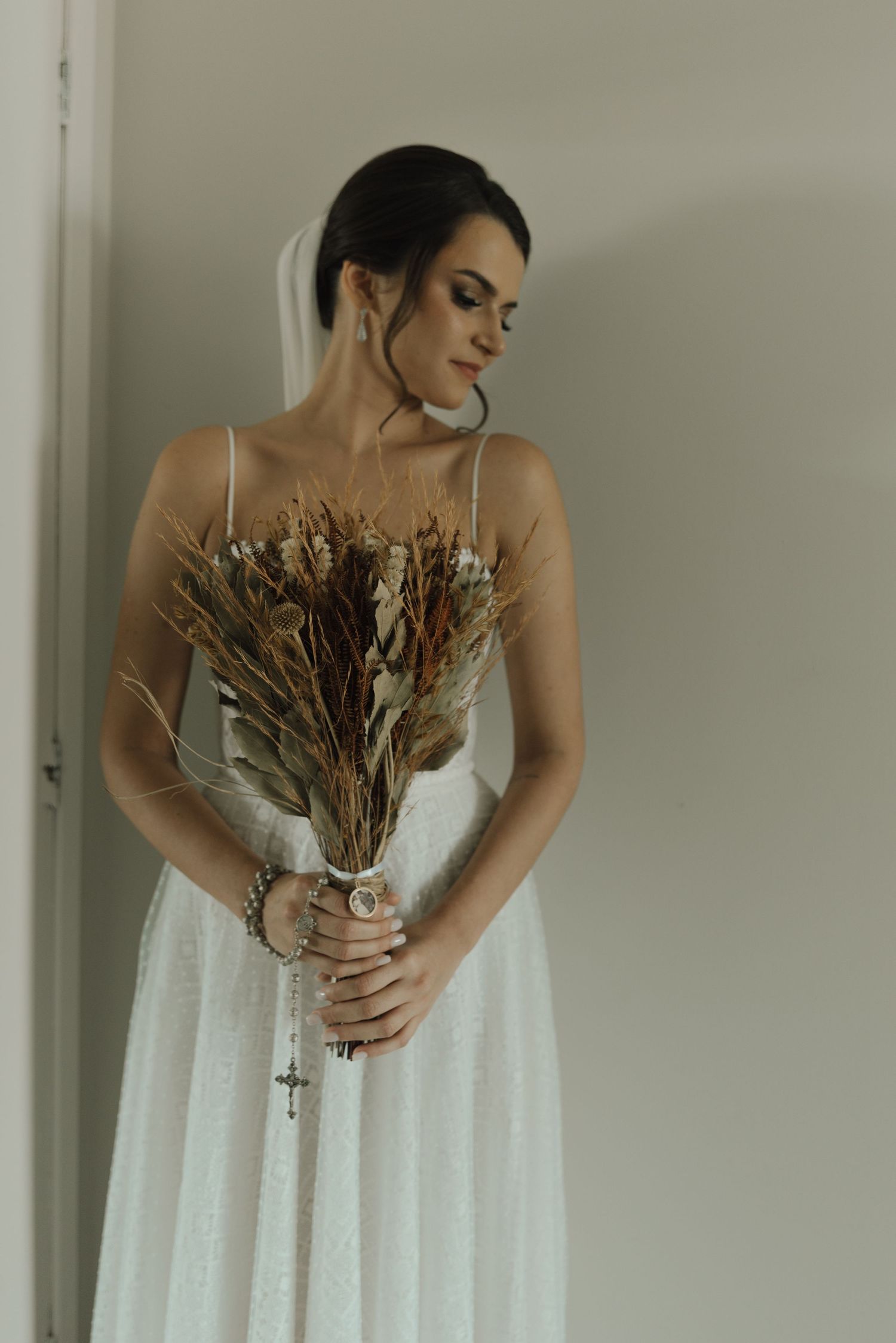 Bride in simple white dress holding dried flower bouquet against neutral wall.
