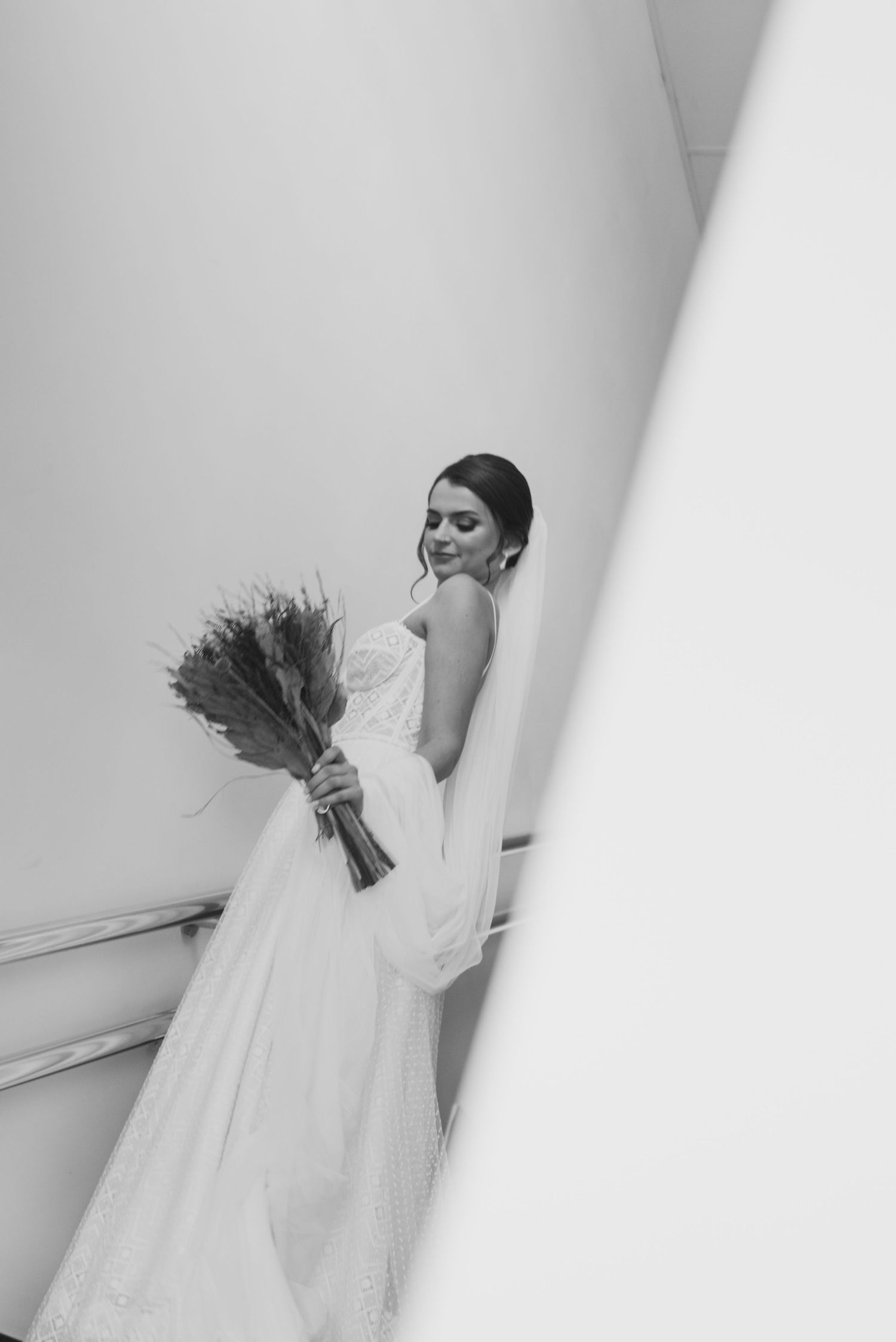 A bride in a white wedding dress poses elegantly with a bouquet against a light colored wall.
