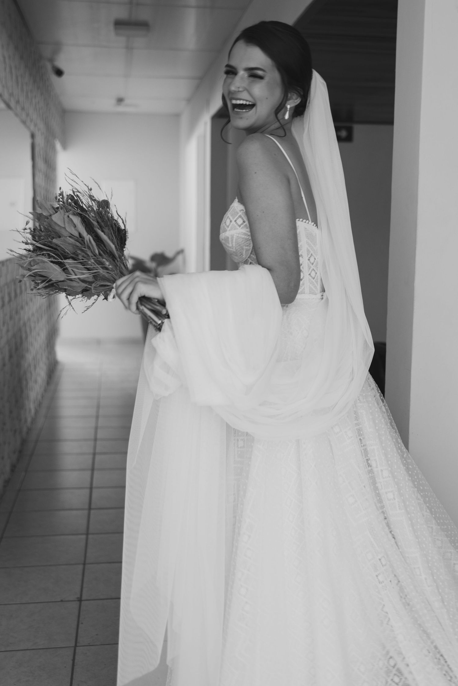 A joyful bride laughs while holding a dried flower bouquet in a hallway.