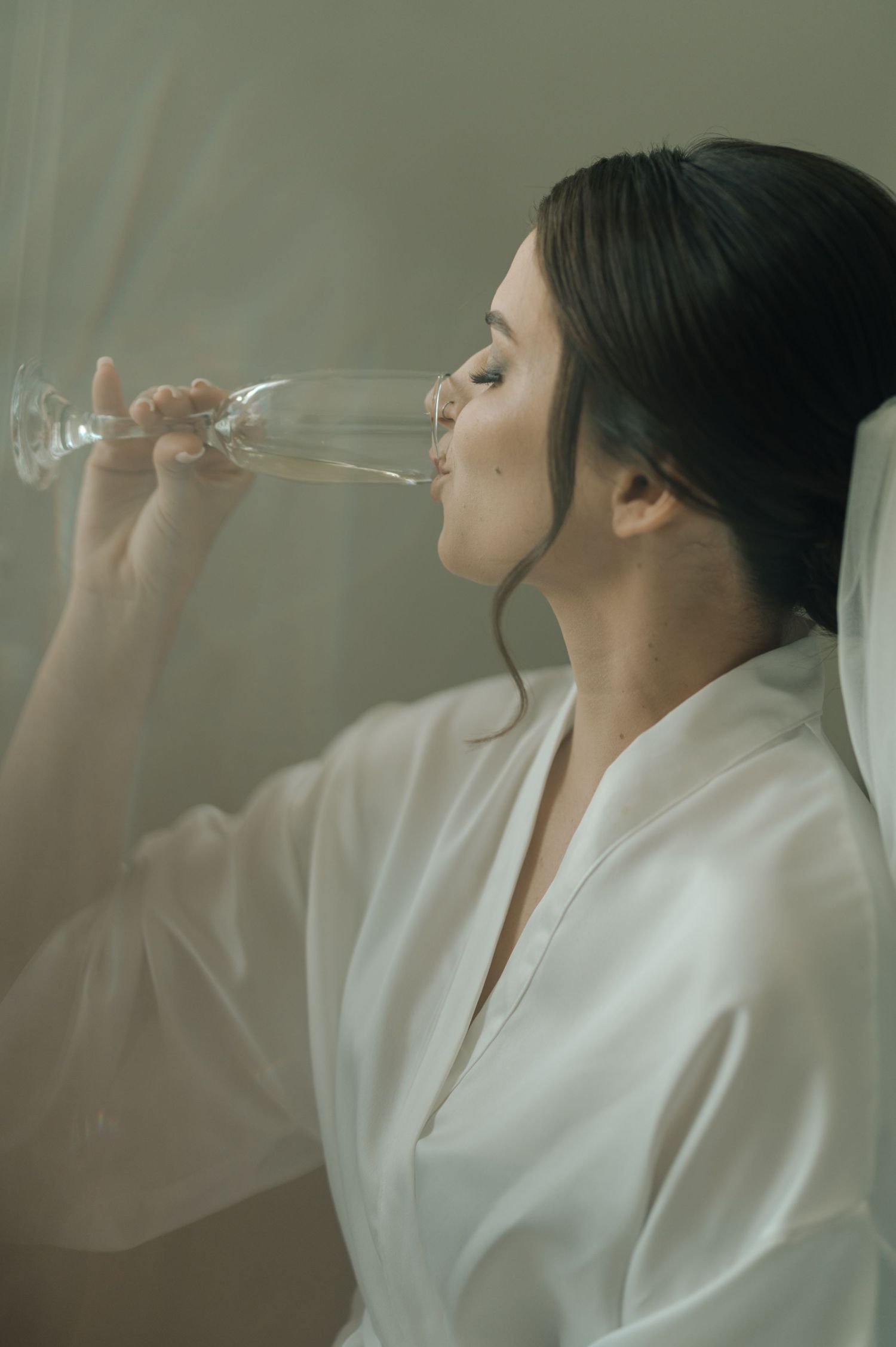 A bride in white robe sips champagne while wearing her wedding veil.