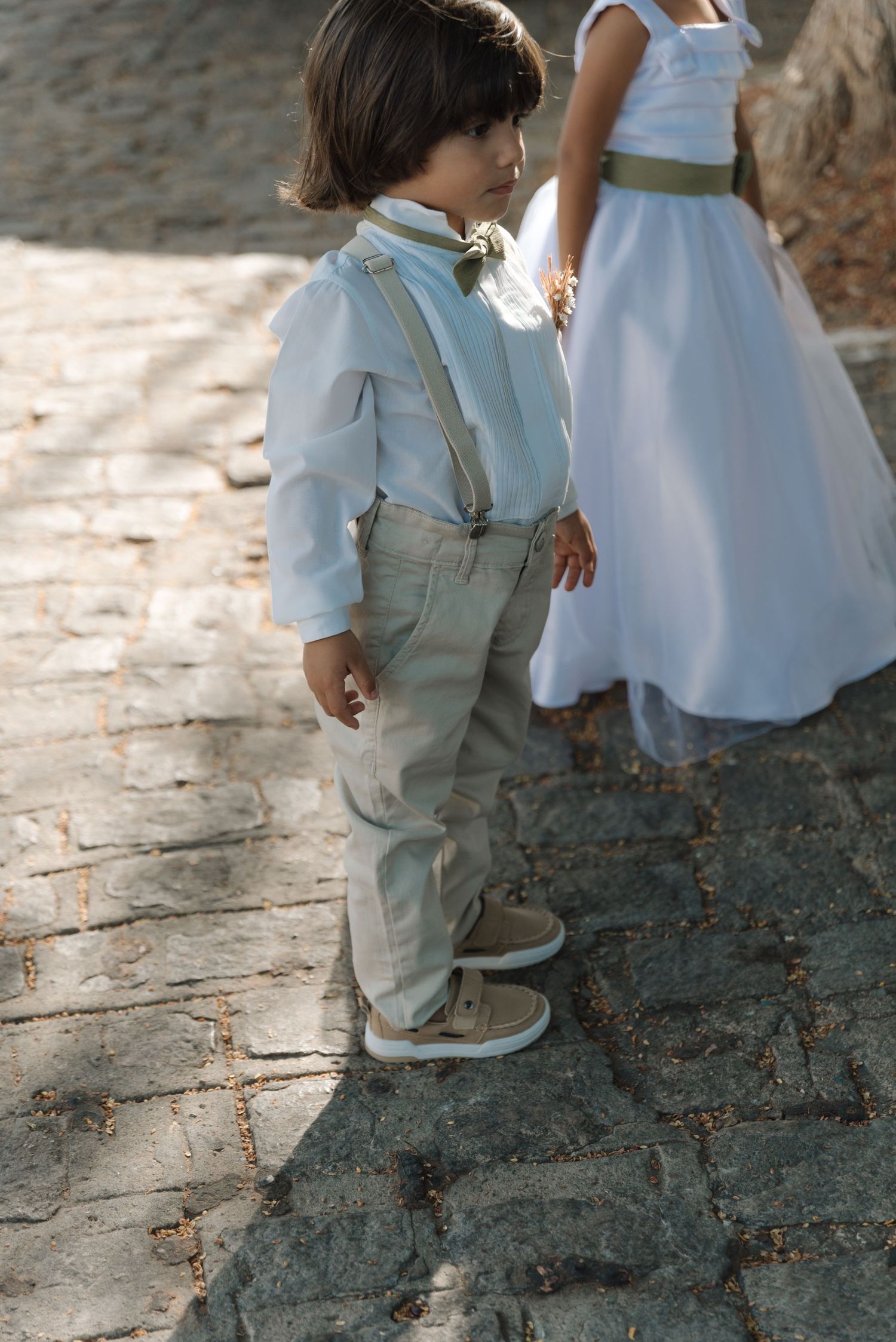 Young ring bearer in light suspender outfit stands on stone path.