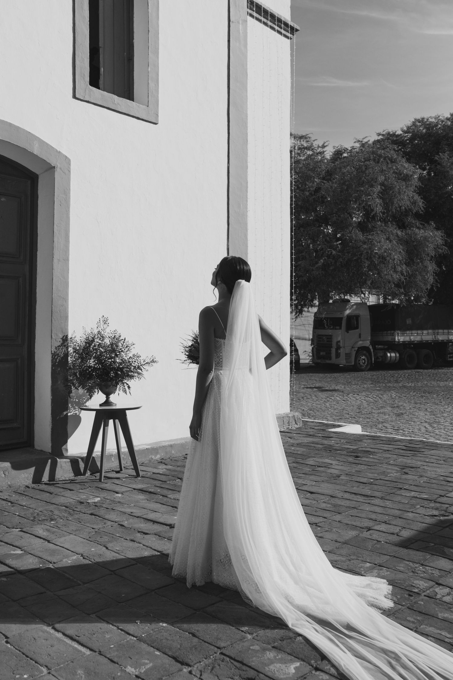 A black and white portrait of a bride standing against a white church wall with trailing veil.