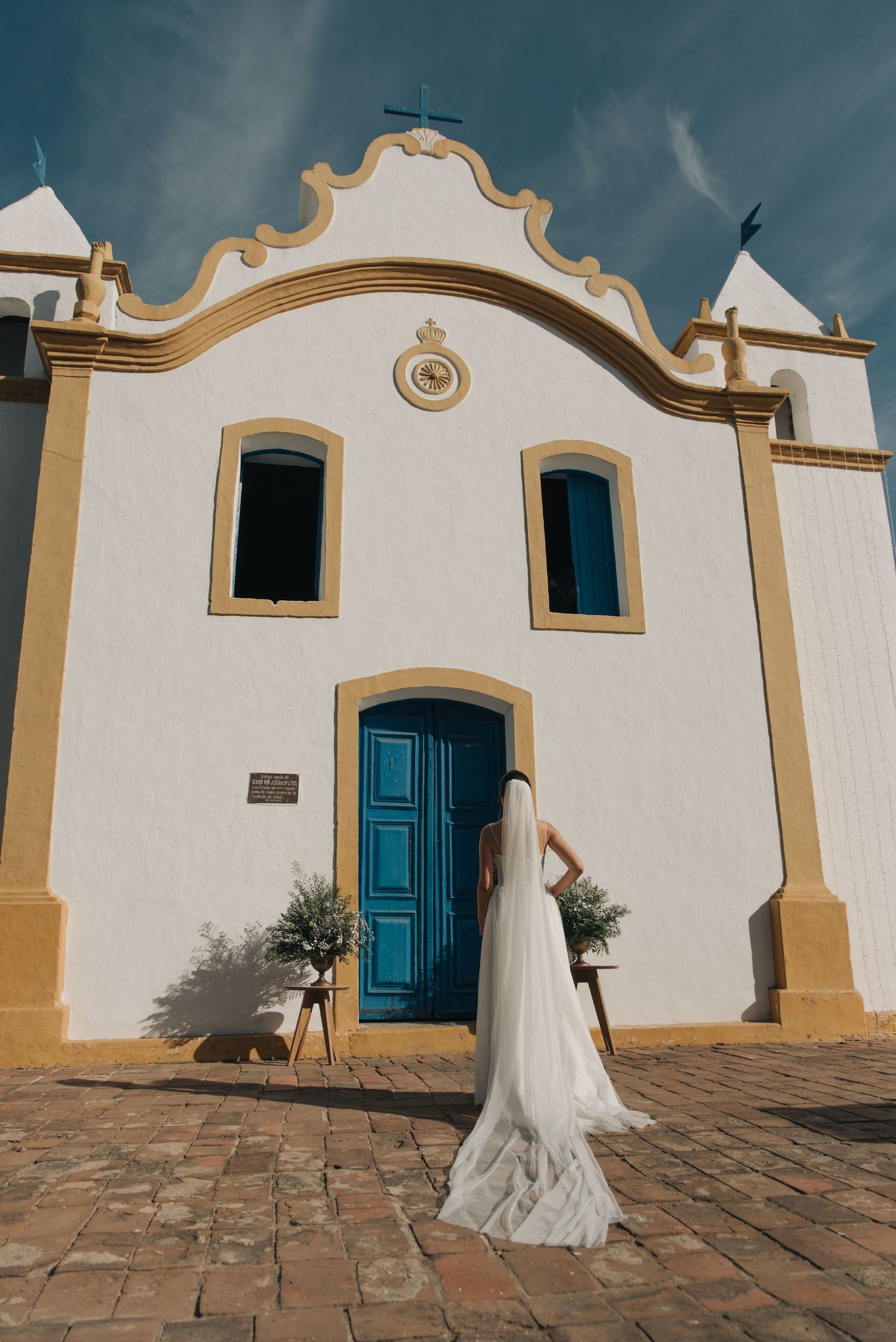 A bride in white dress poses in front of a Portuguese colonial church with blue door and gold trim.
