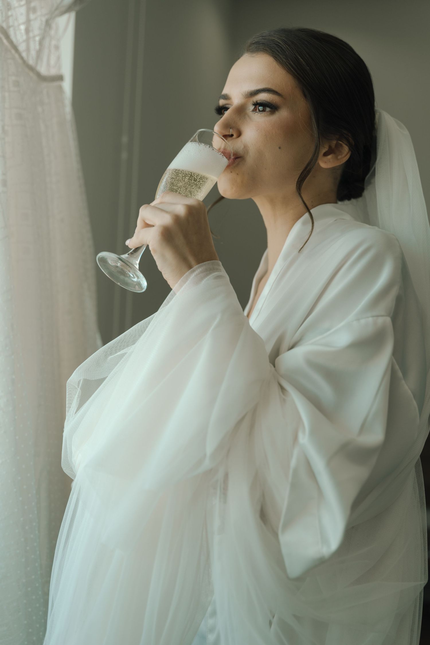 A bride in white robe enjoys champagne while standing by a wedding dress.