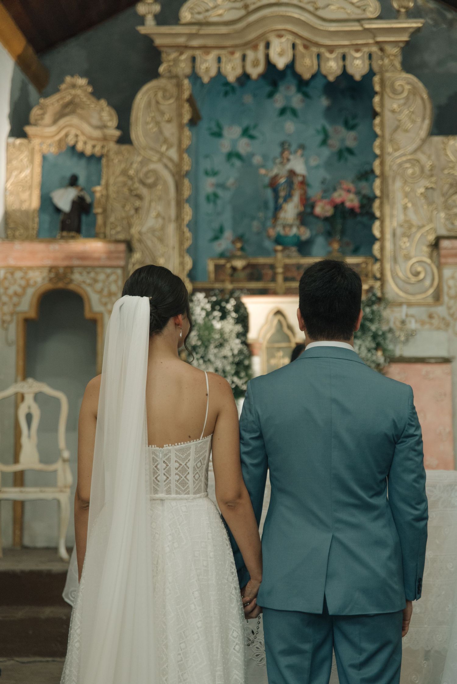 A couple stands before an ornate church altar with turquoise and gold baroque details during their wedding ceremony.