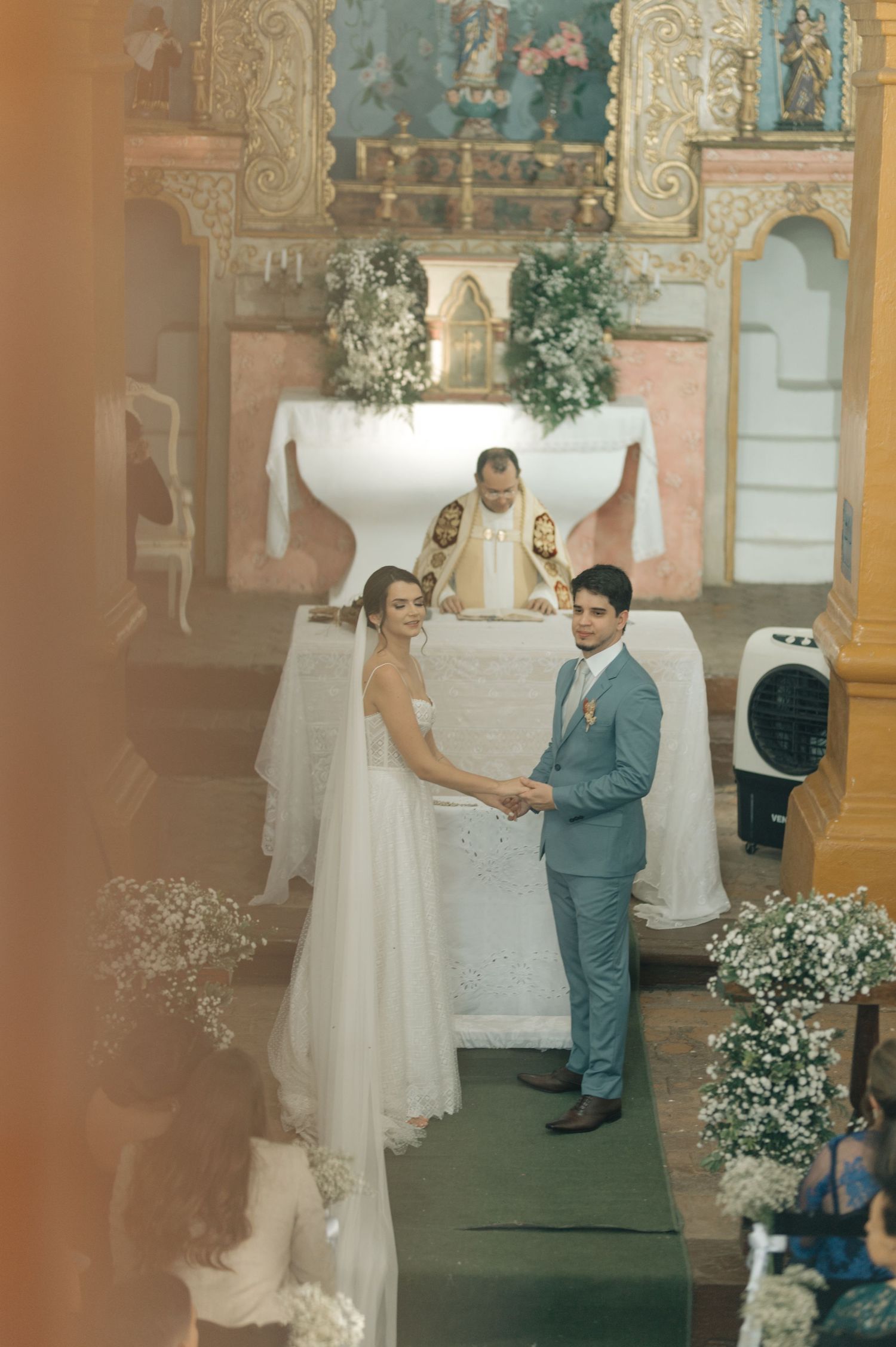 A wedding ceremony at an ornate church altar with floral arrangements and a couple exchanging vows.