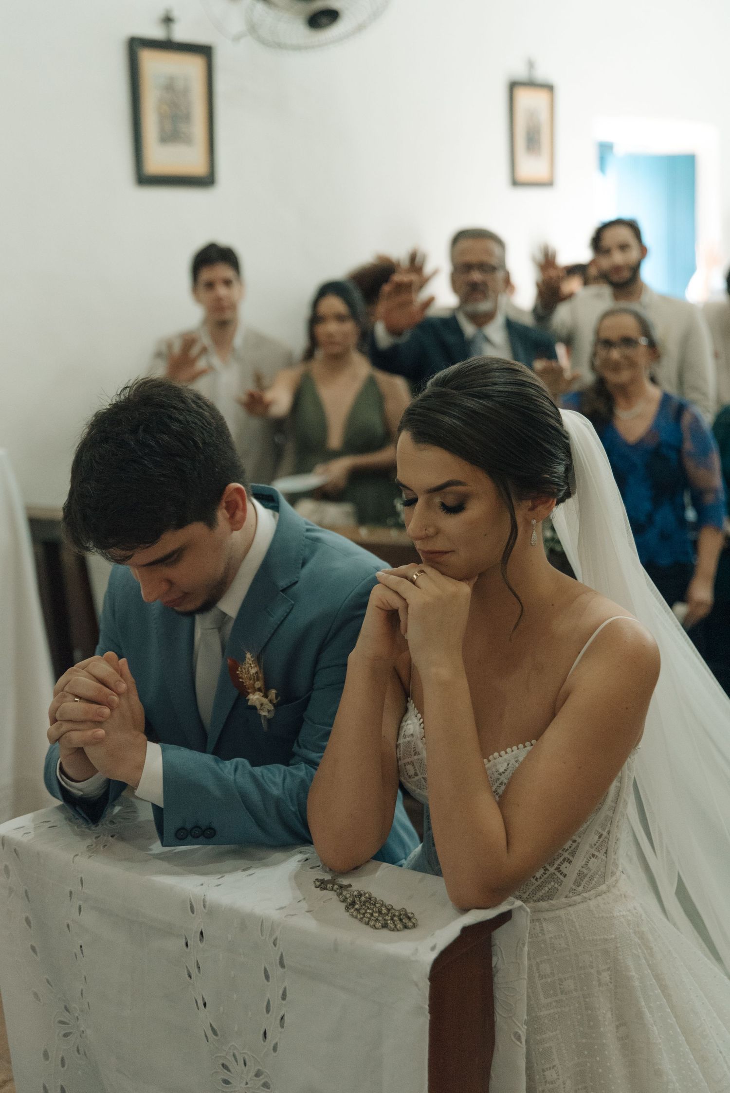 A couple praying together during their wedding ceremony while guests wave in the background.
