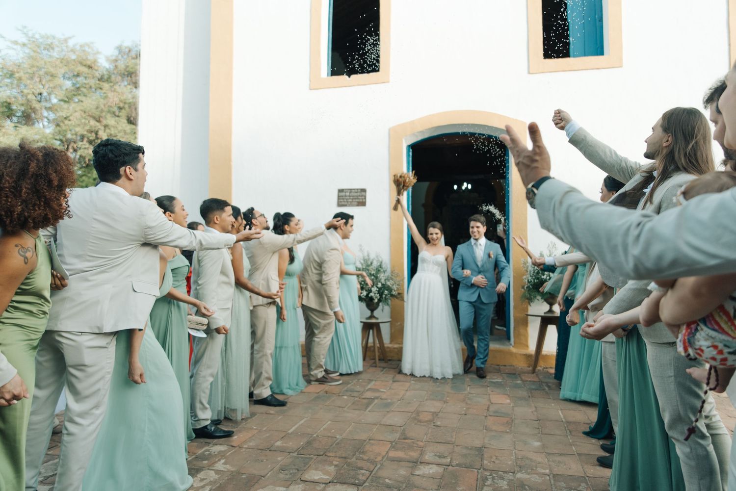 Wedding guests in mint green and beige attire form an aisle outside a white church.