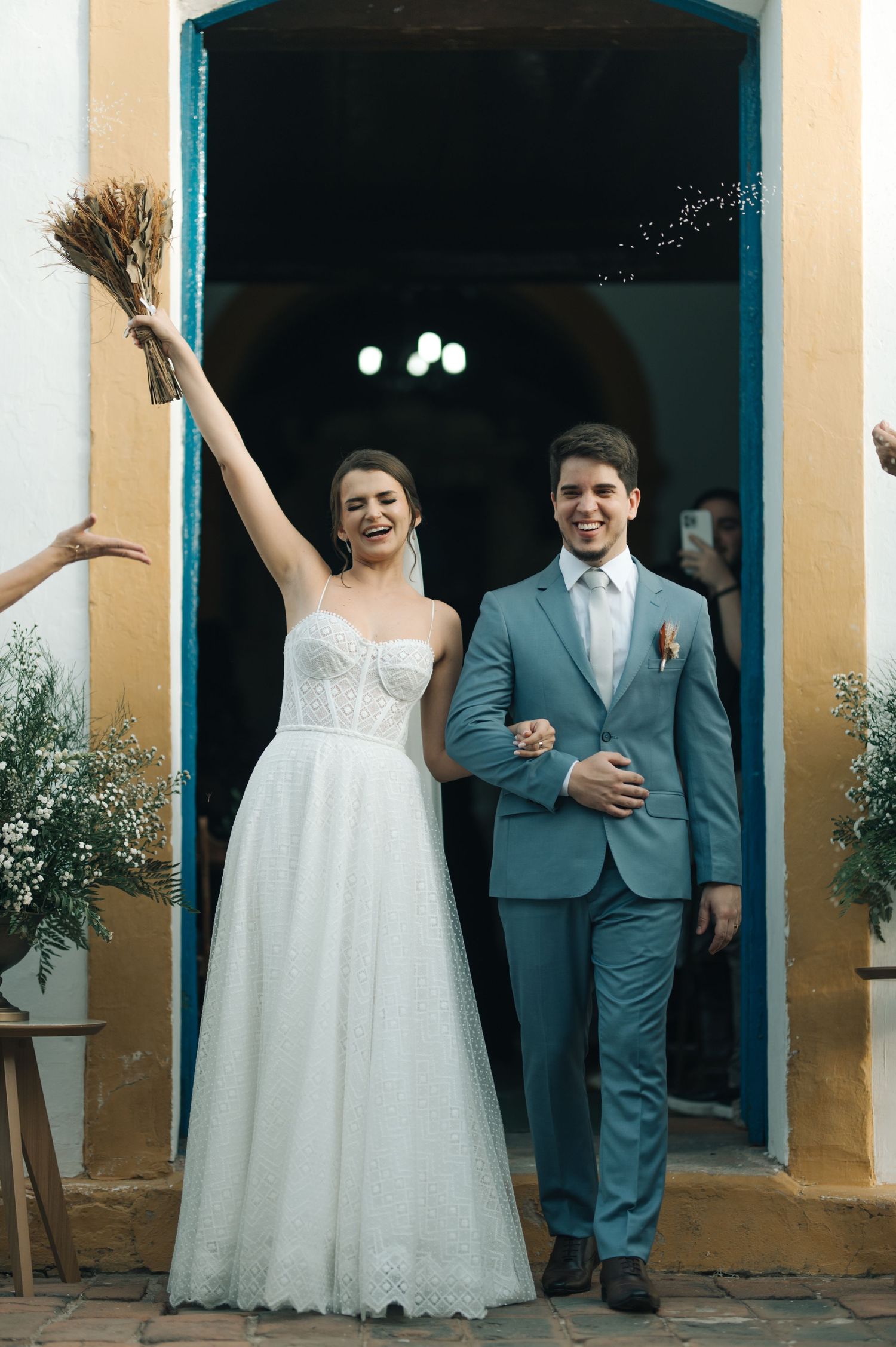 Newlyweds exit church in celebration with bride in white gown and groom in light blue suit.