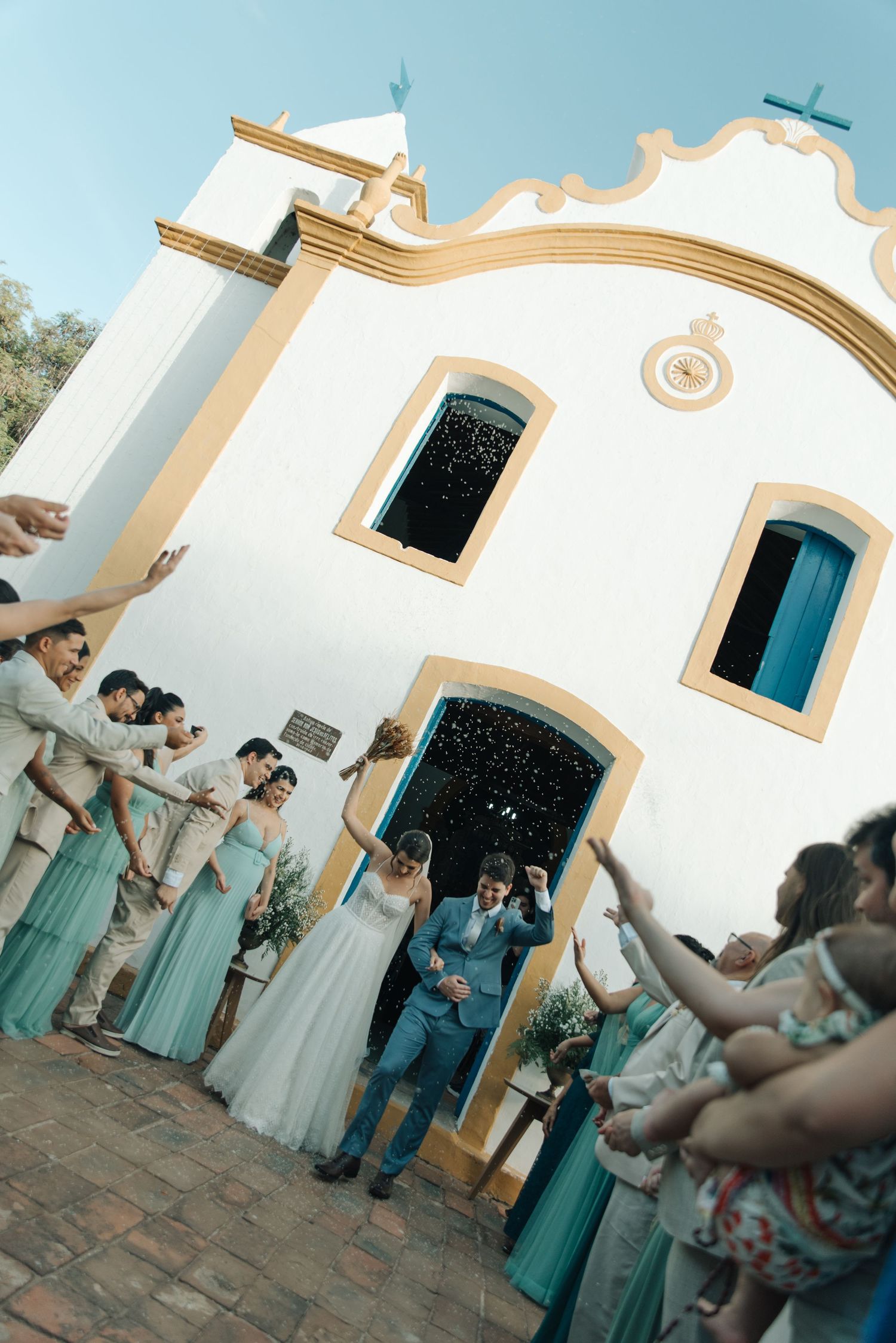 Wedding party cheers as newlyweds exit white colonial-style church building.
