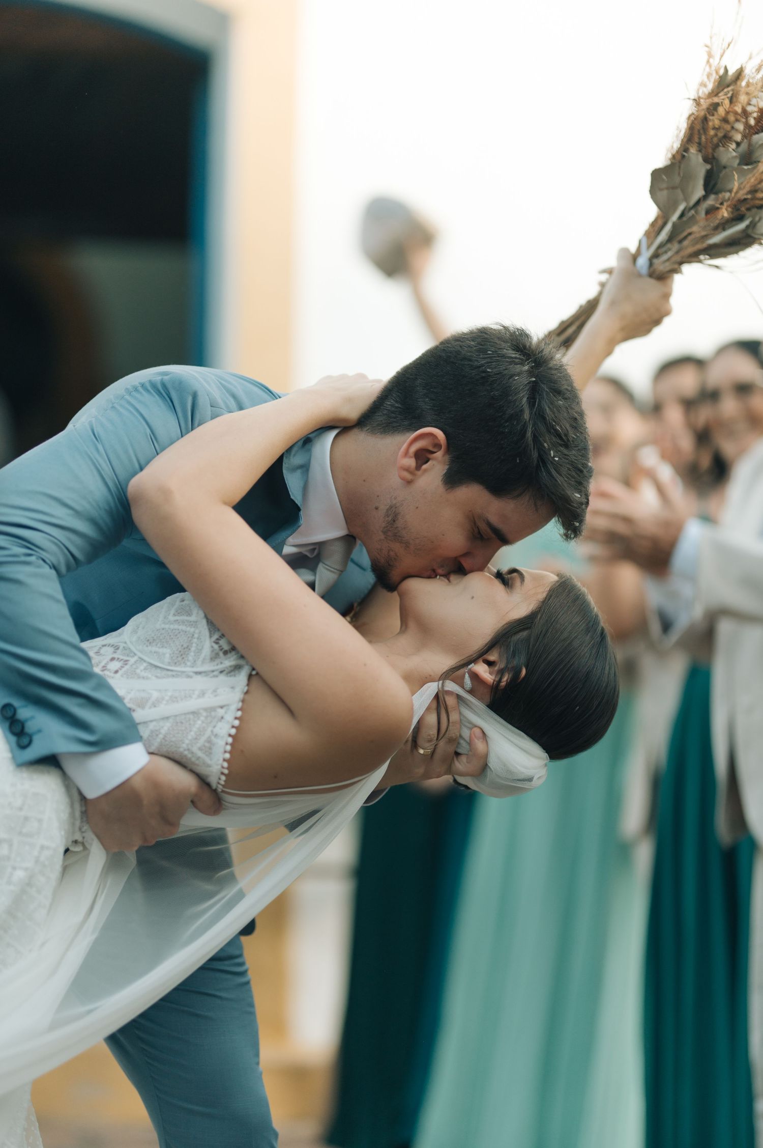 Romantic dip kiss between newlyweds outside church while guests celebrate.