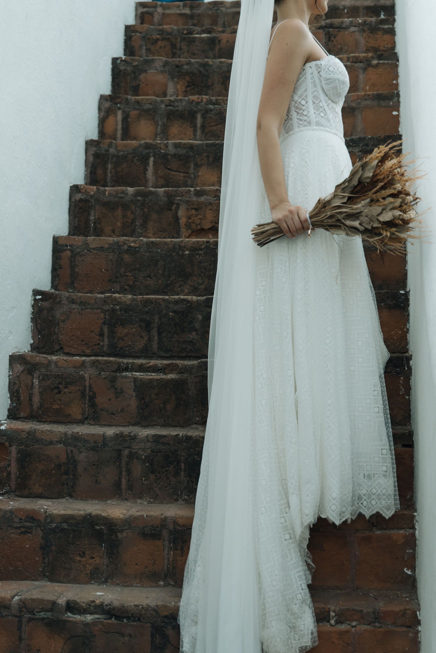 Bride in a lace white dress holding dried flowers stands beside rustic brick stairs.
