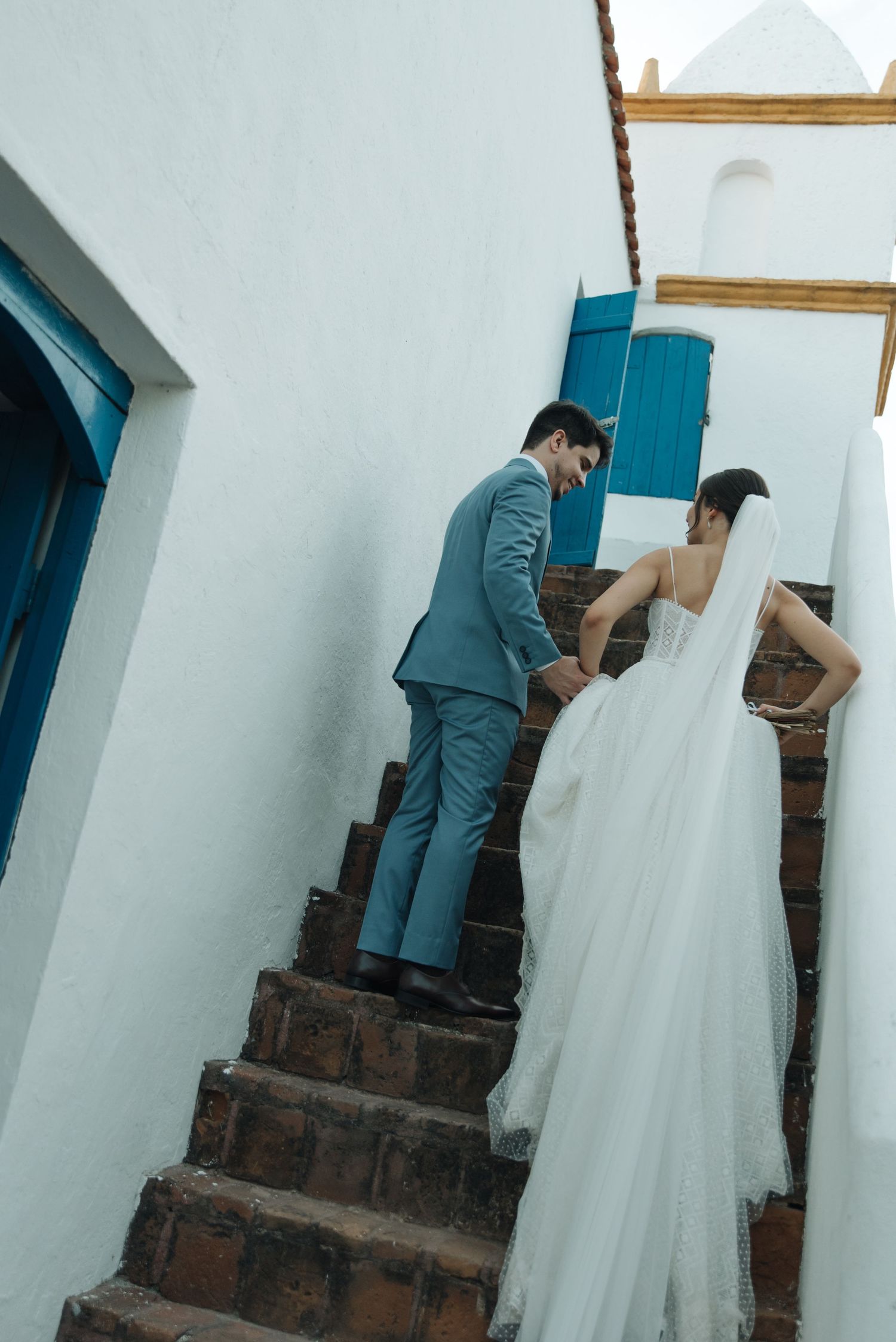 Wedding couple navigating brick stairs against white walls with blue shutters.