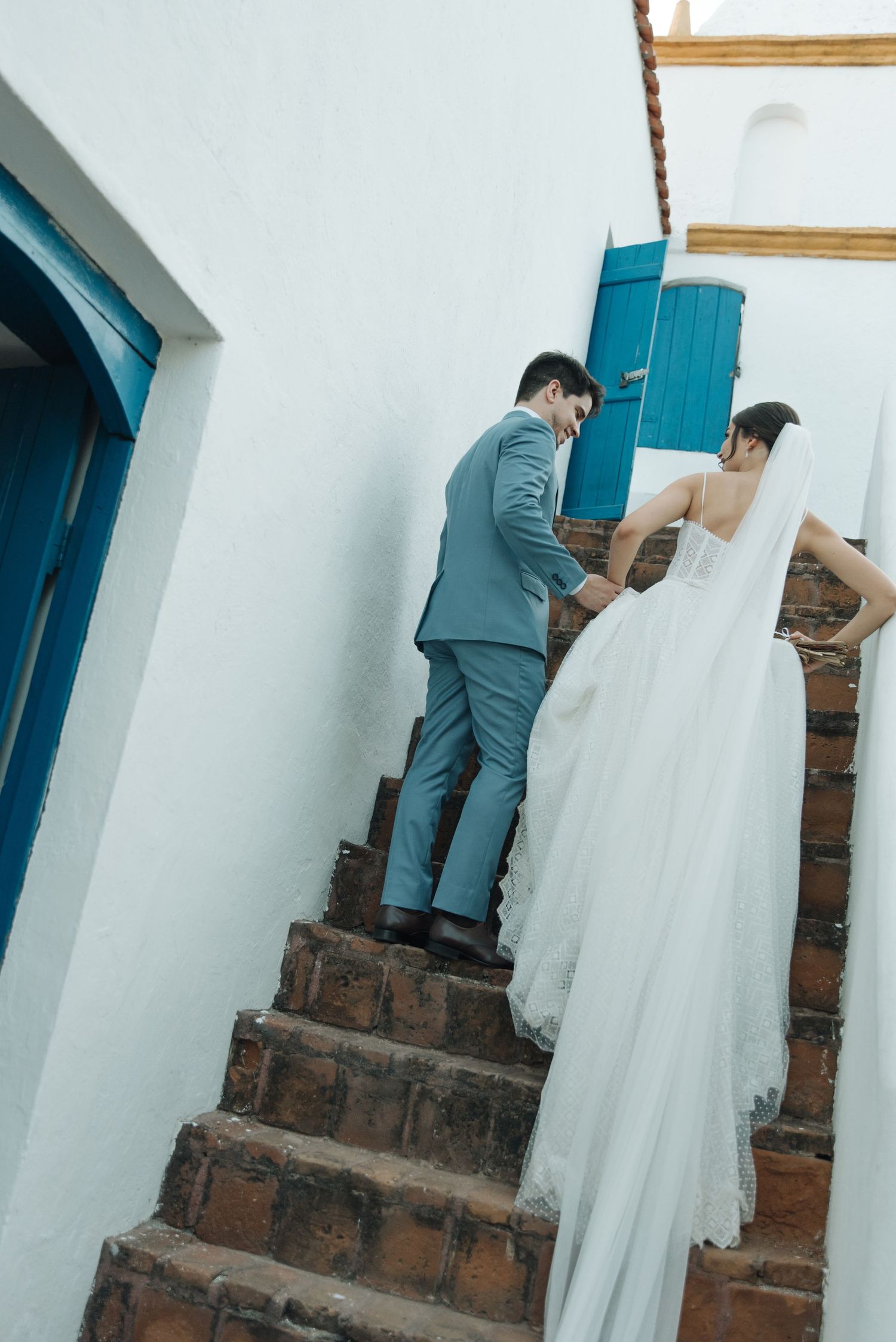 Newlyweds climbing rustic brick steps with Mediterranean-style architecture in background.