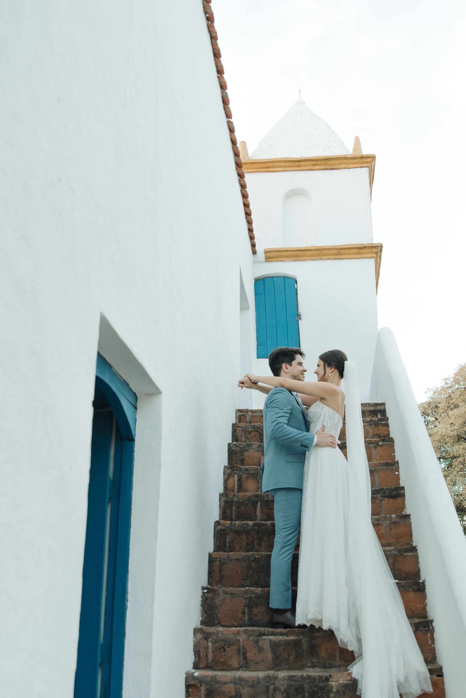 Newlyweds embrace on brick staircase against white Mediterranean building with blue shutters.