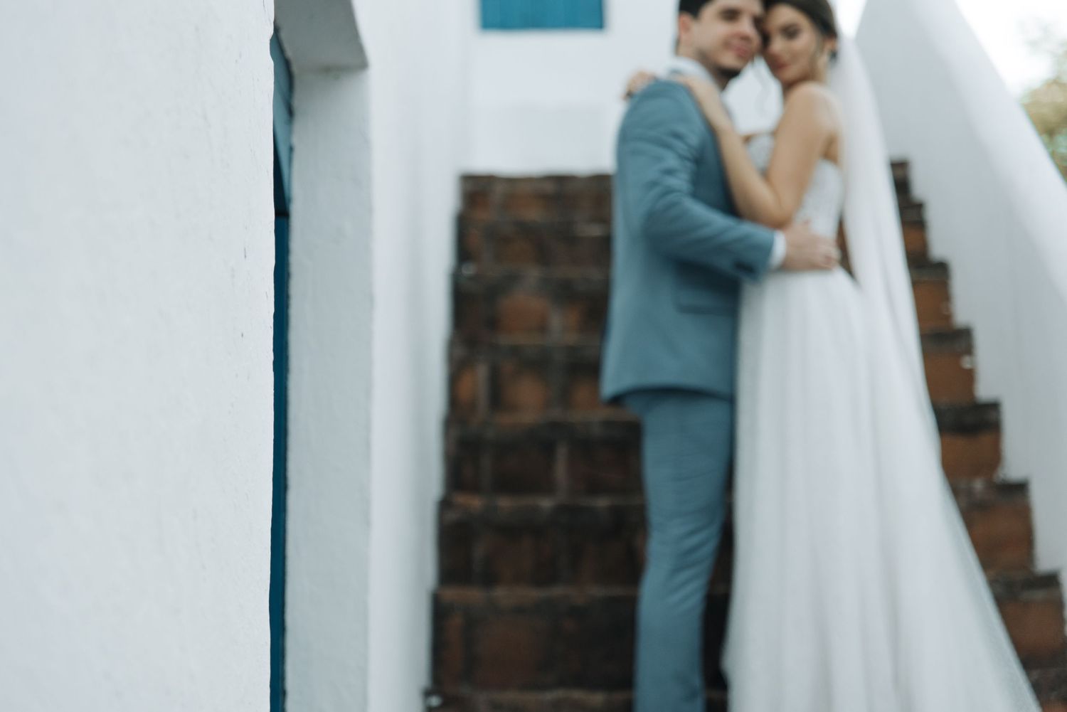 A romantic wedding couple embraces on rustic stone steps against a white Mediterranean style building.