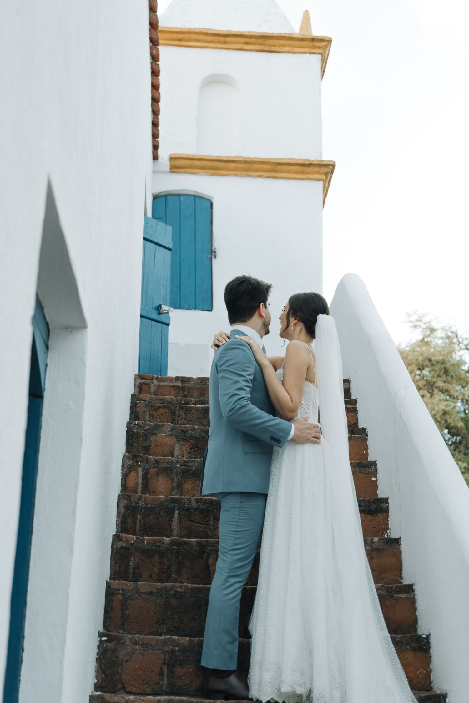 Newlyweds share an intimate moment on stone stairs with turquoise shutters and white walls.
