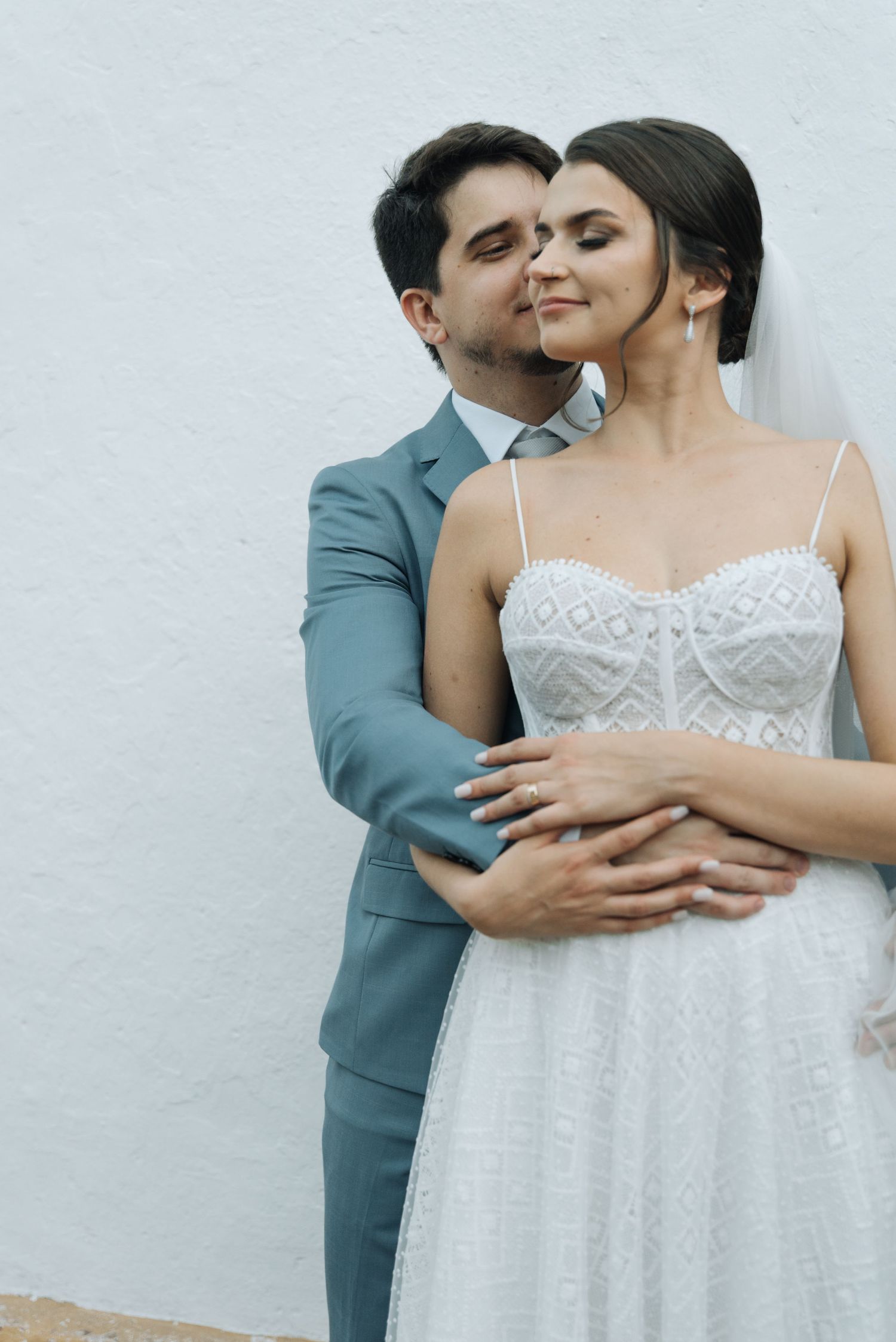 A tender moment between bride in lace dress and groom in blue suit against white wall.
