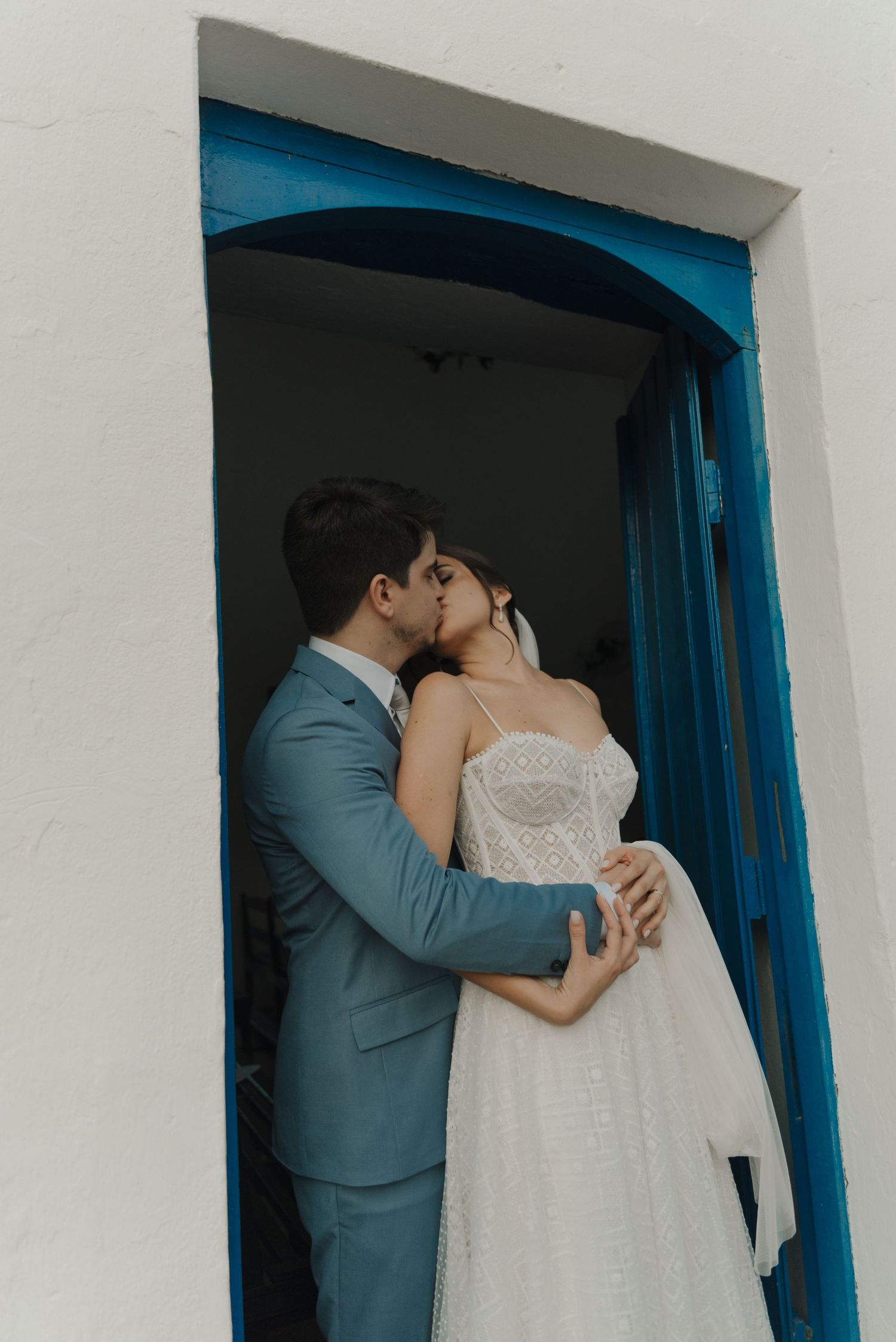 Intimate kiss between newlyweds in blue-trimmed doorway of Mediterranean building.