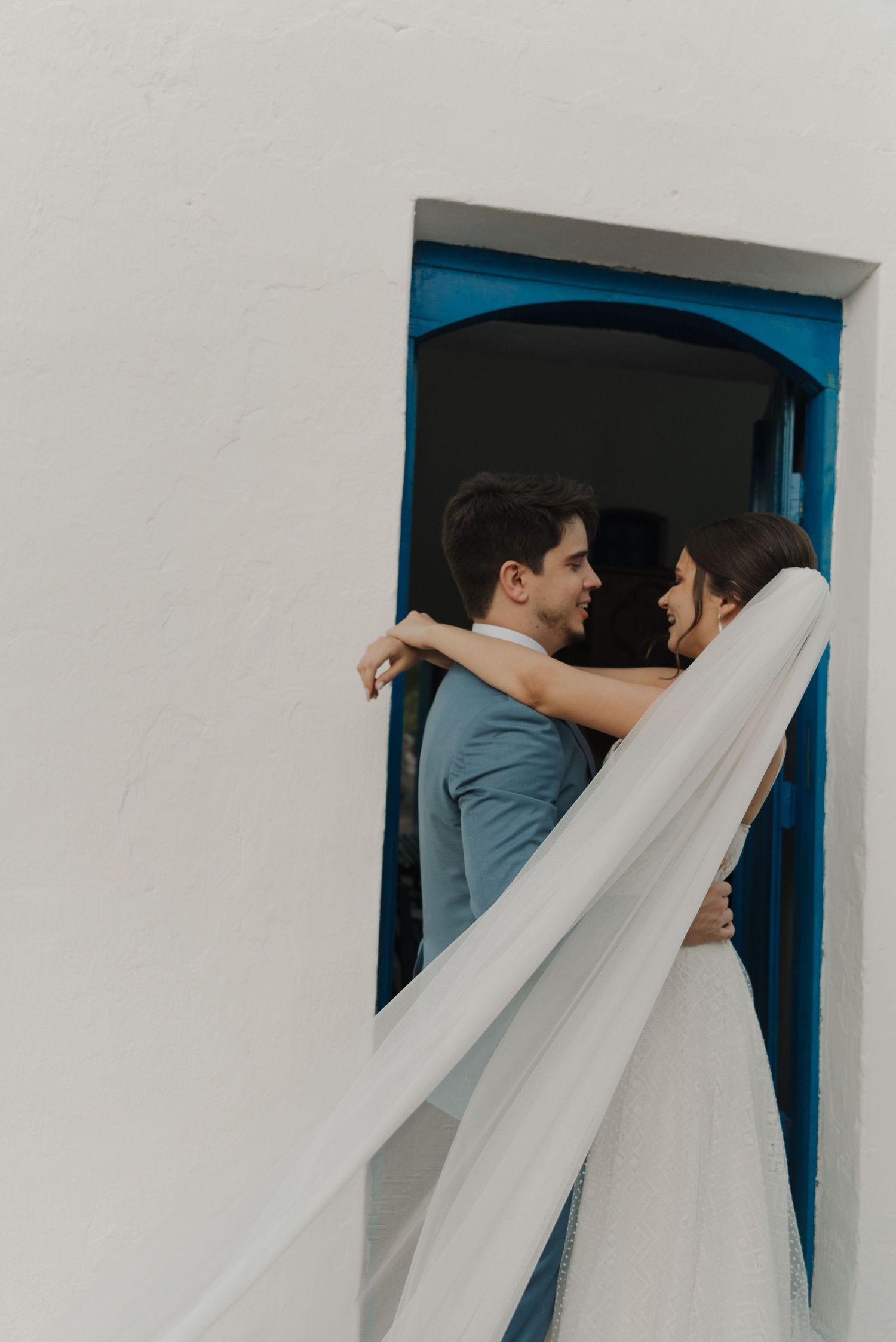 Bride and groom share embrace in blue-trimmed doorway with flowing white veil.