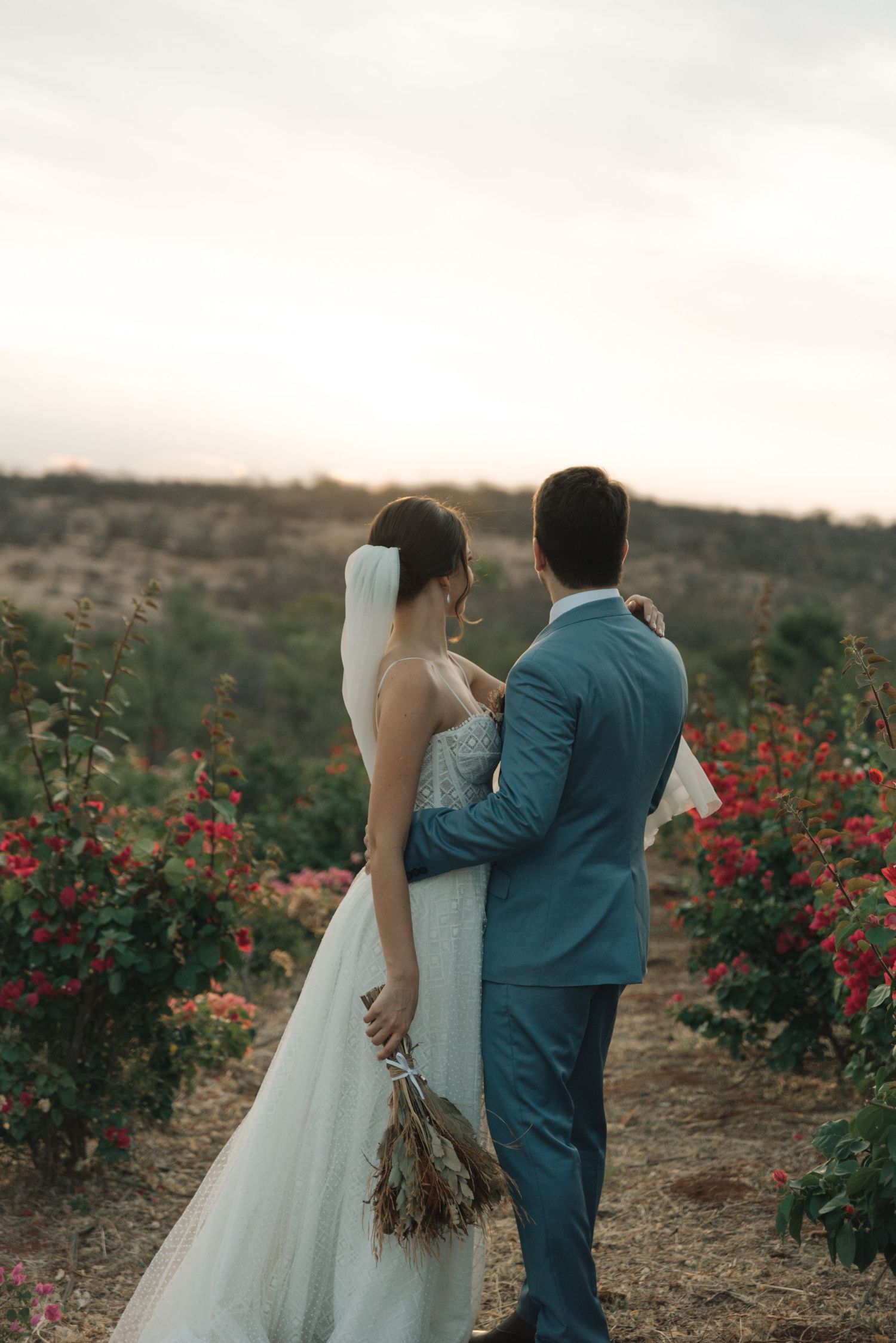 A romantic wedding couple embraces near red flowers in a desert landscape at sunset.