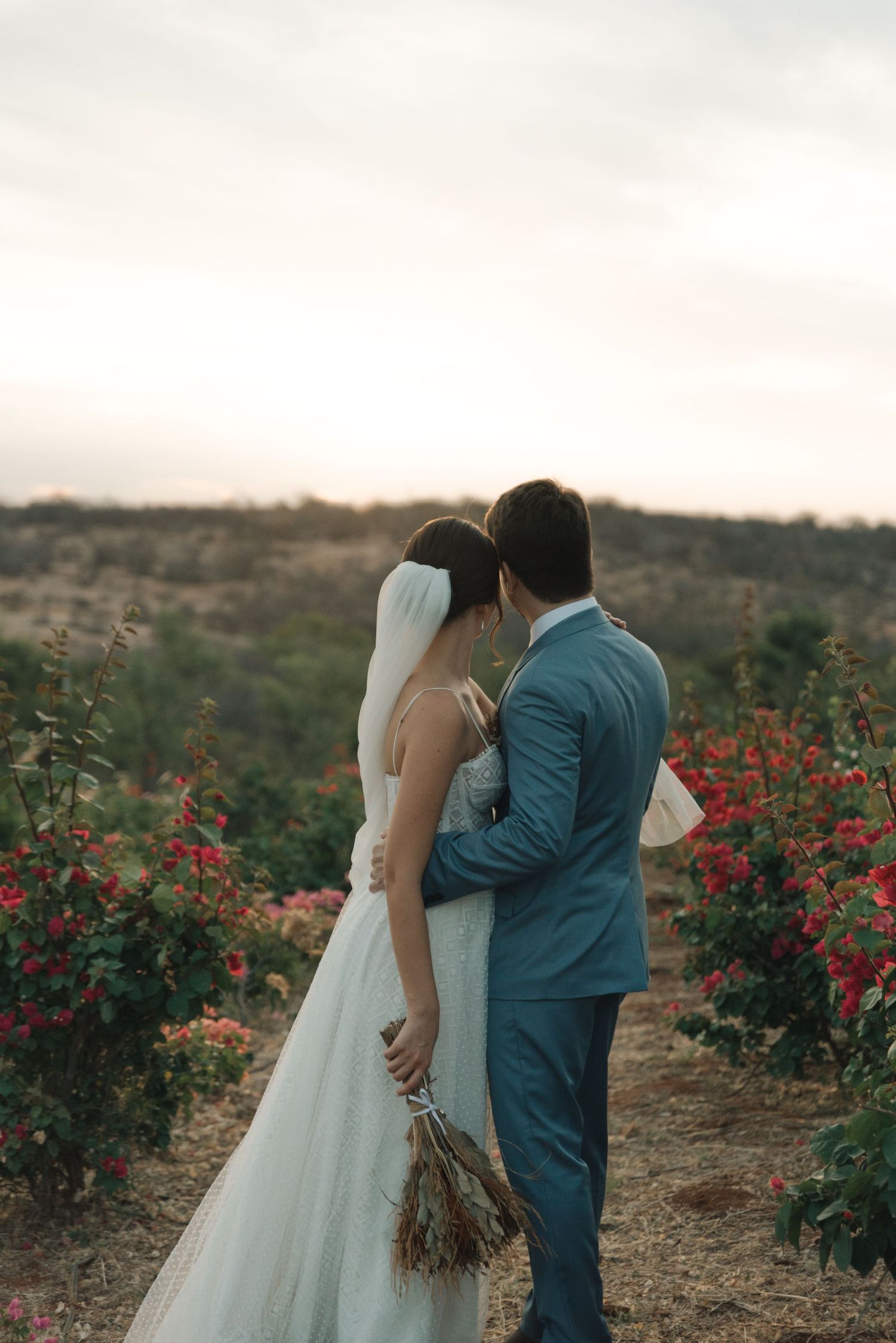 Newlyweds share an intimate moment among blooming red flowers in a rustic outdoor setting.