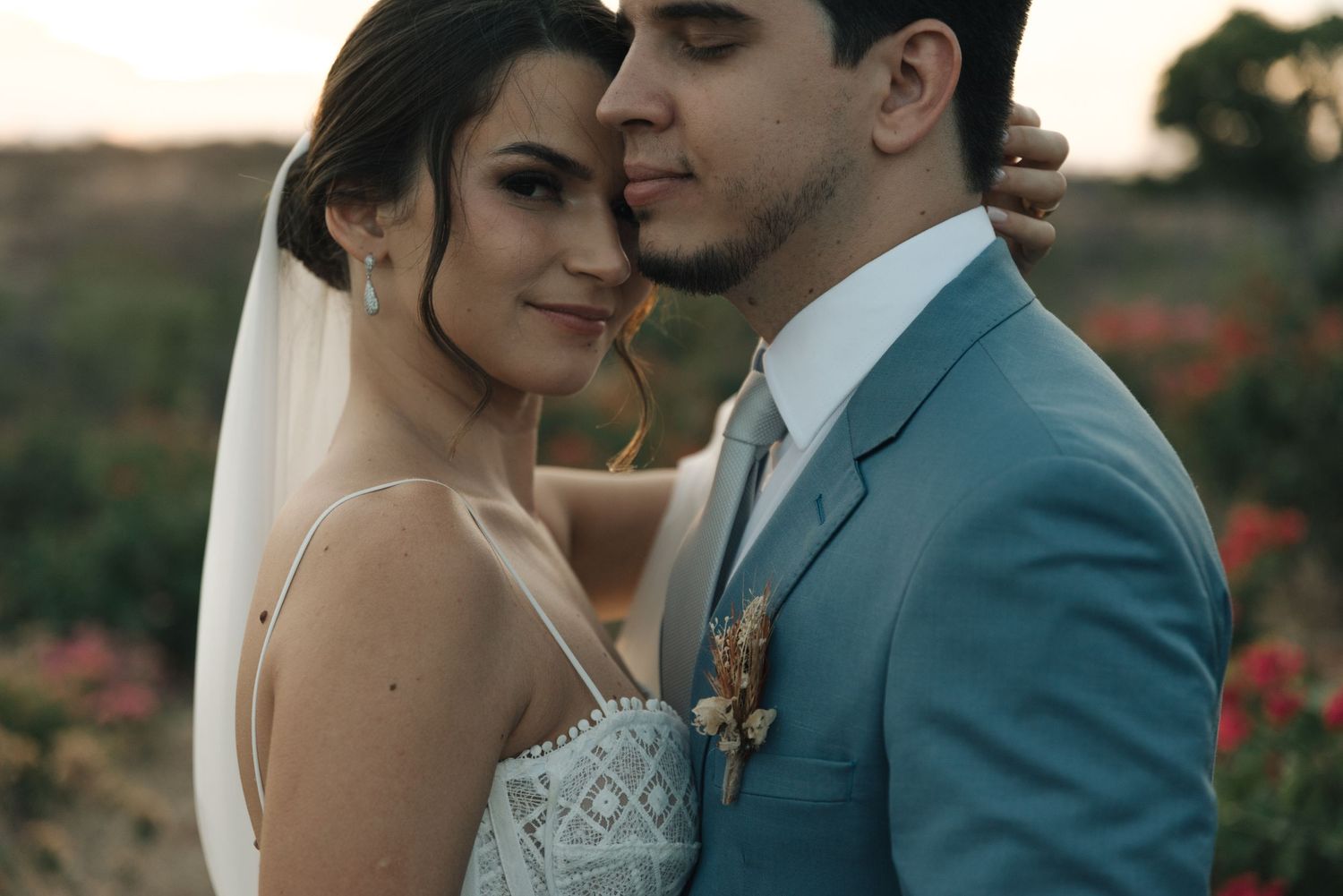 Bride in lace dress and groom in blue suit share an intimate moment in a desert landscape.