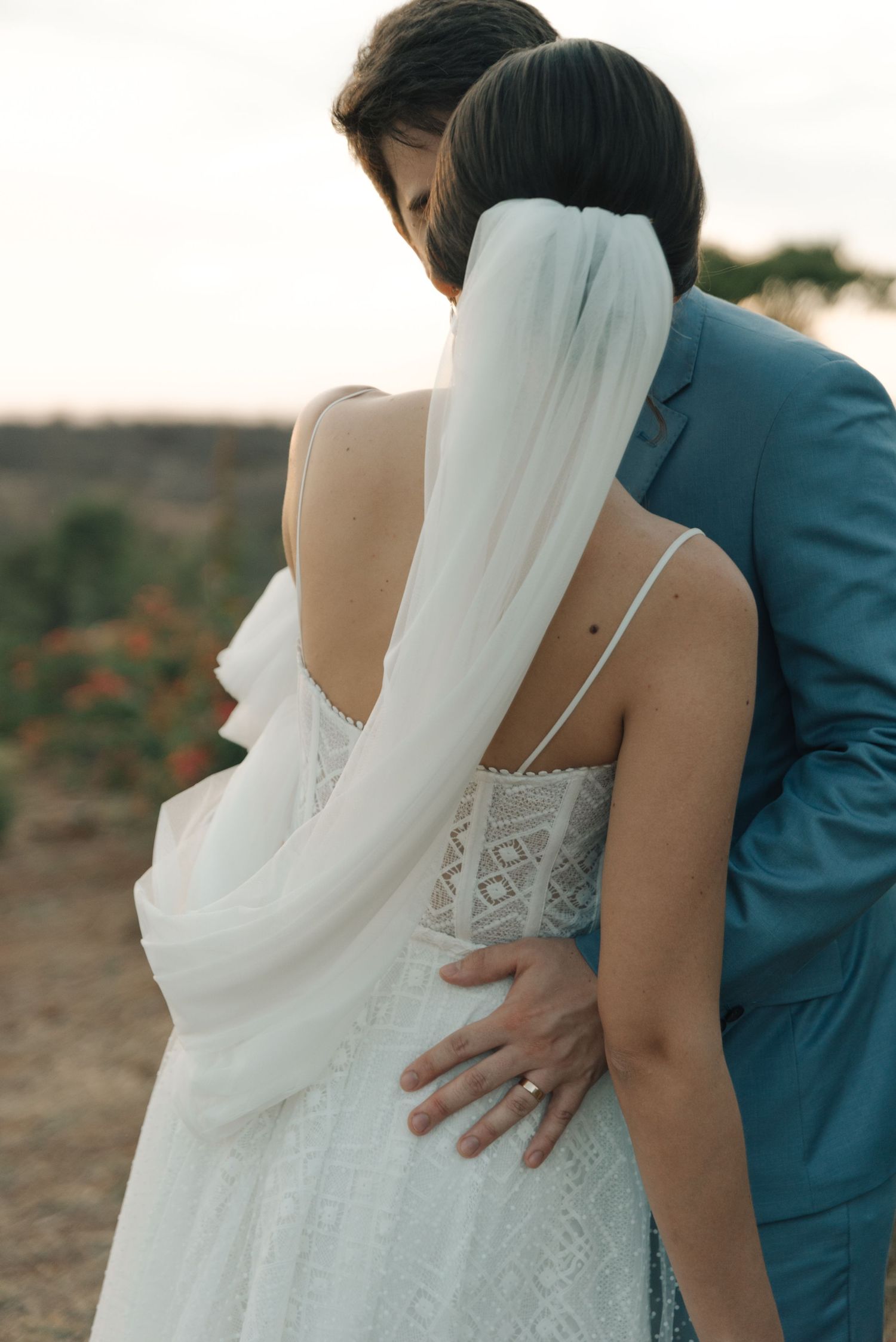 Close-up of bride's detailed lace dress and flowing veil as couple embraces outdoors.