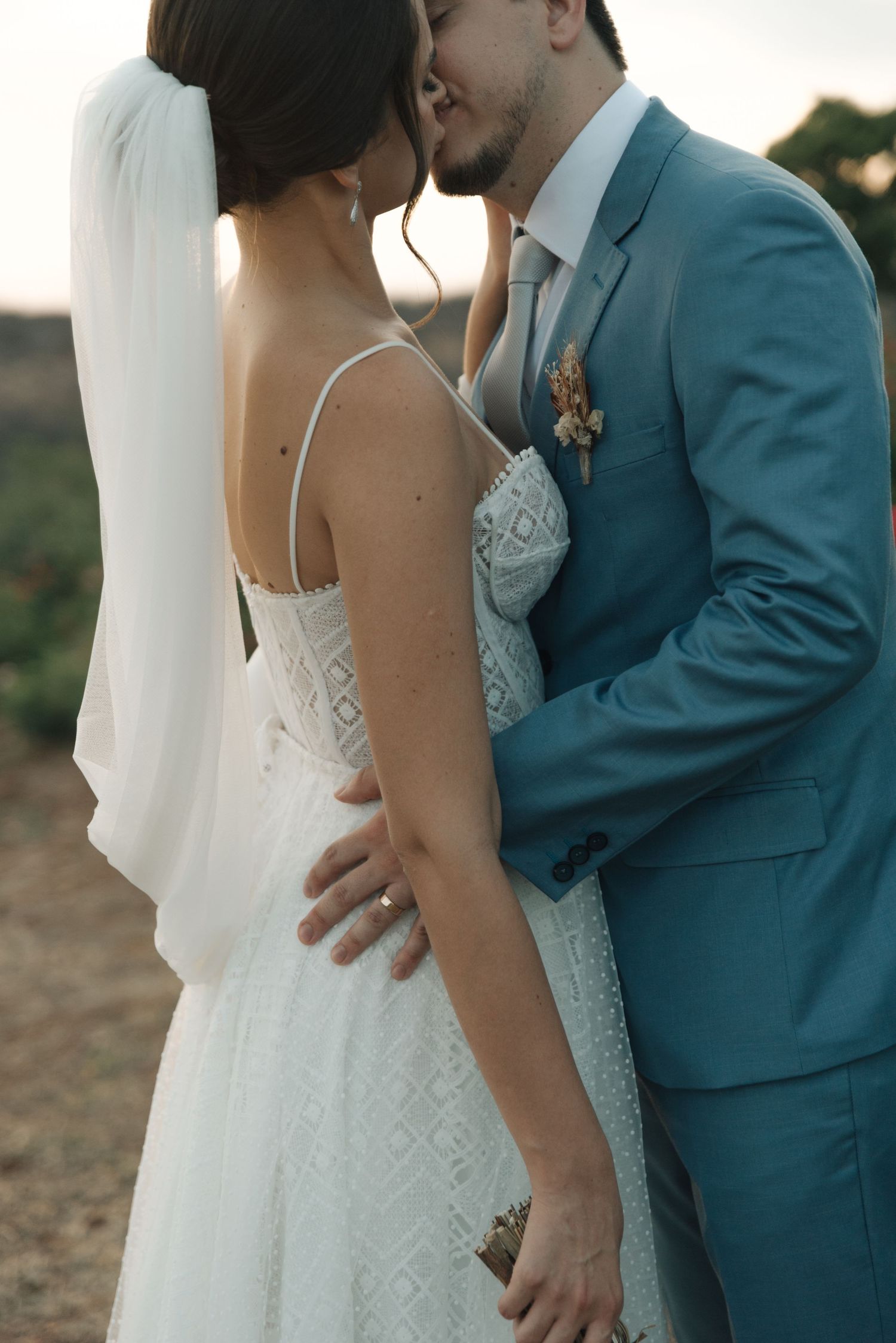 Newlyweds share an intimate moment outdoors, with the bride in a white lace gown and groom in a teal blue suit.