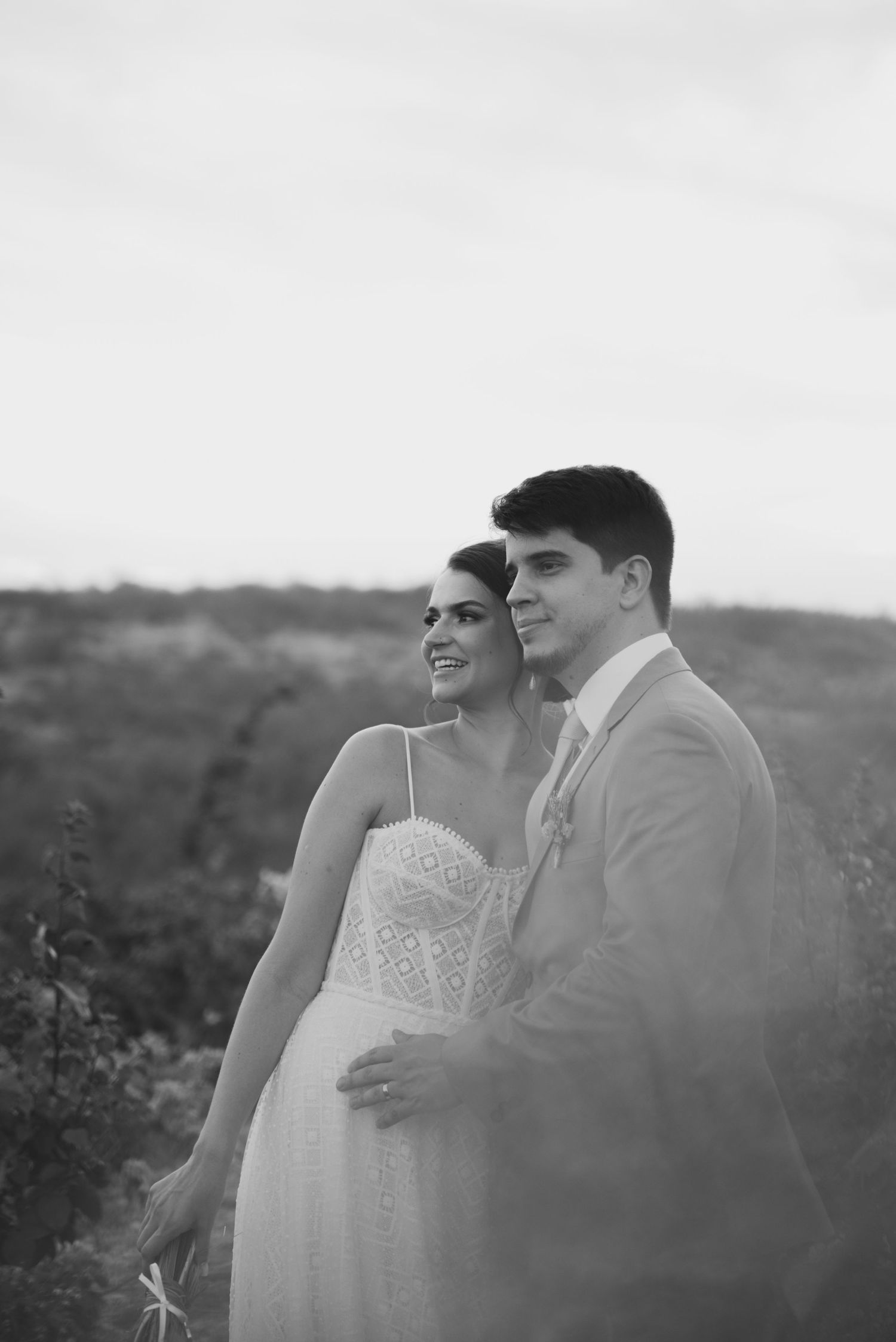 Black and white portrait of a happy couple in wedding attire standing together in a natural outdoor setting.