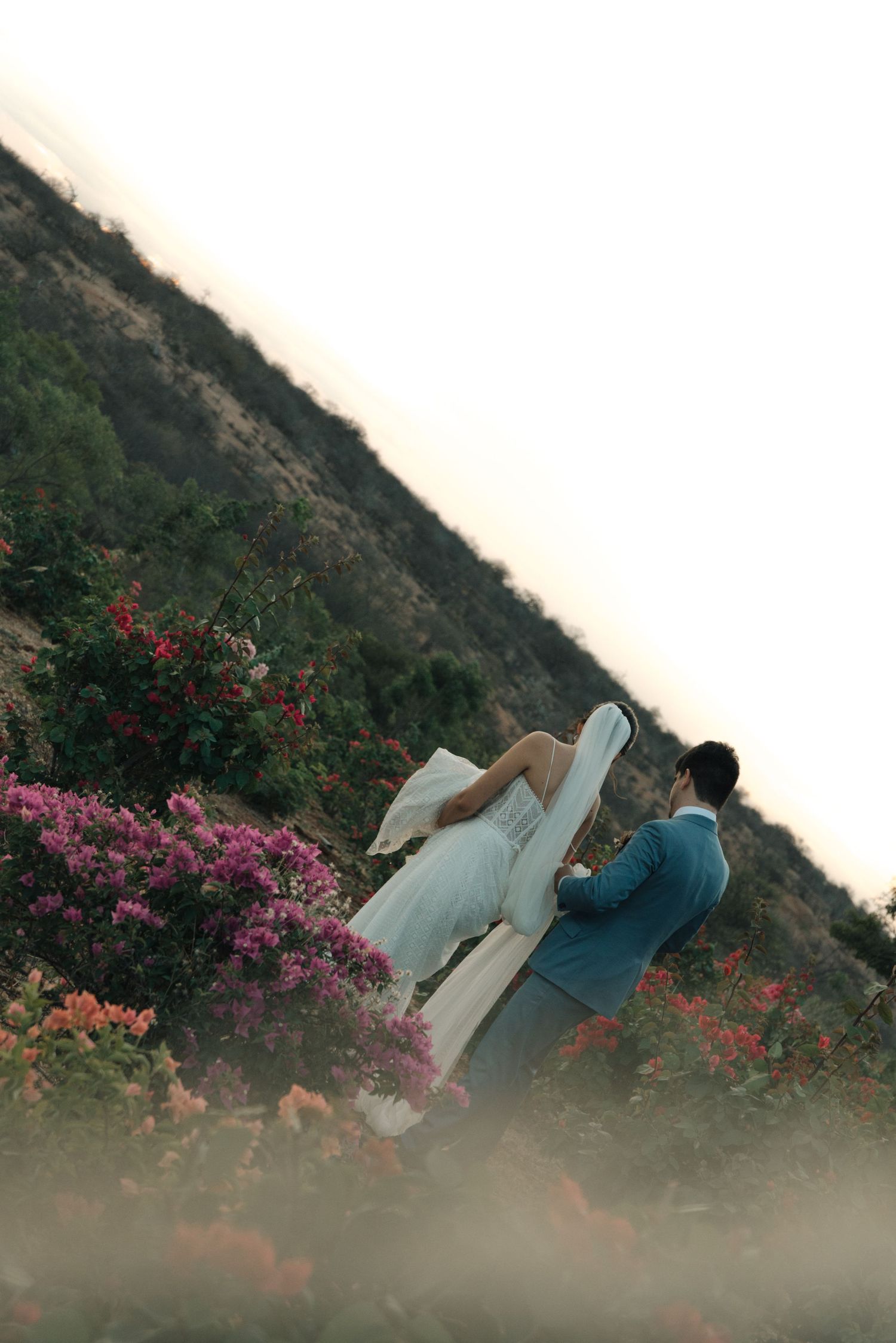 Romantic wedding photo of couple surrounded by colorful flowers on a hillside at sunset.
