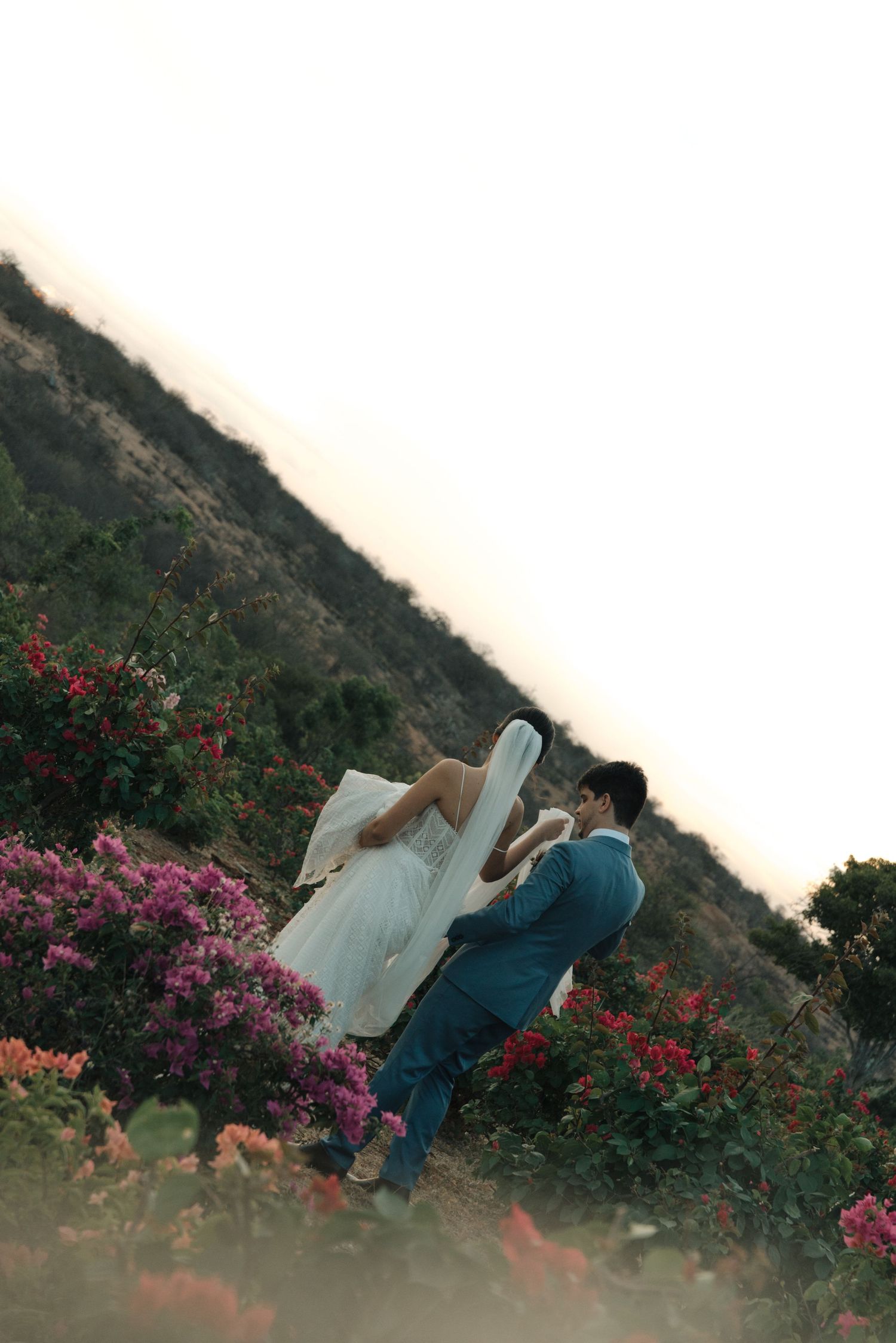 Wedding couple embracing among vibrant pink and red flowers on a scenic hillside.