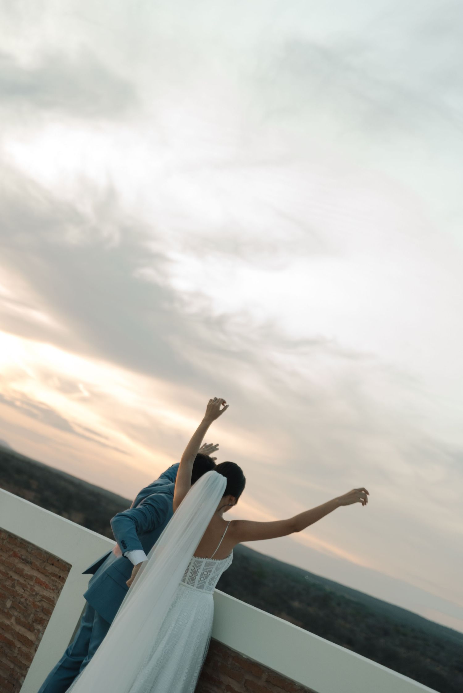 Bride and groom posing playfully at sunset with arms raised against dramatic sky.