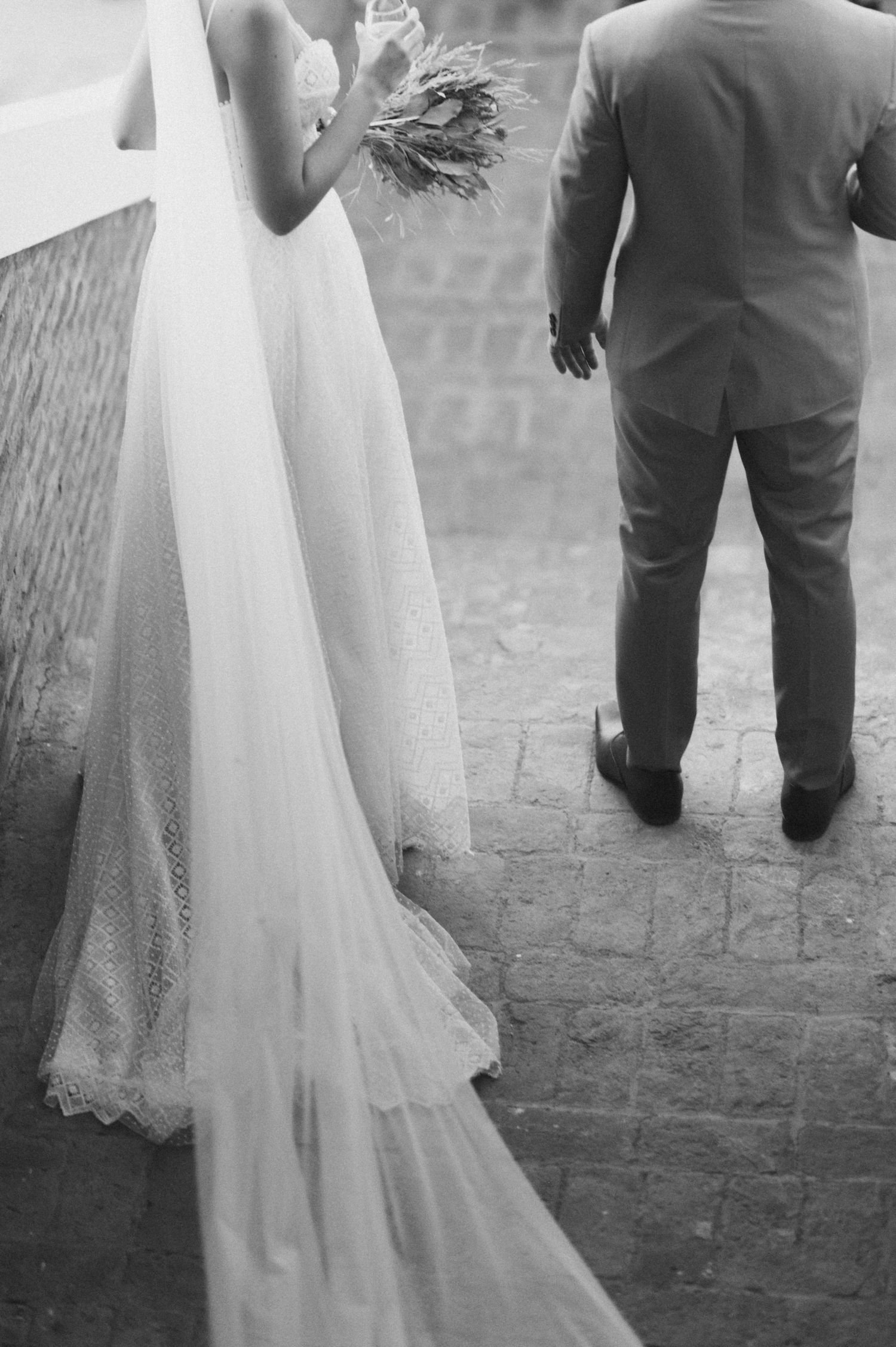 Black and white photo of bride's flowing dress train and groom on stone pathway.