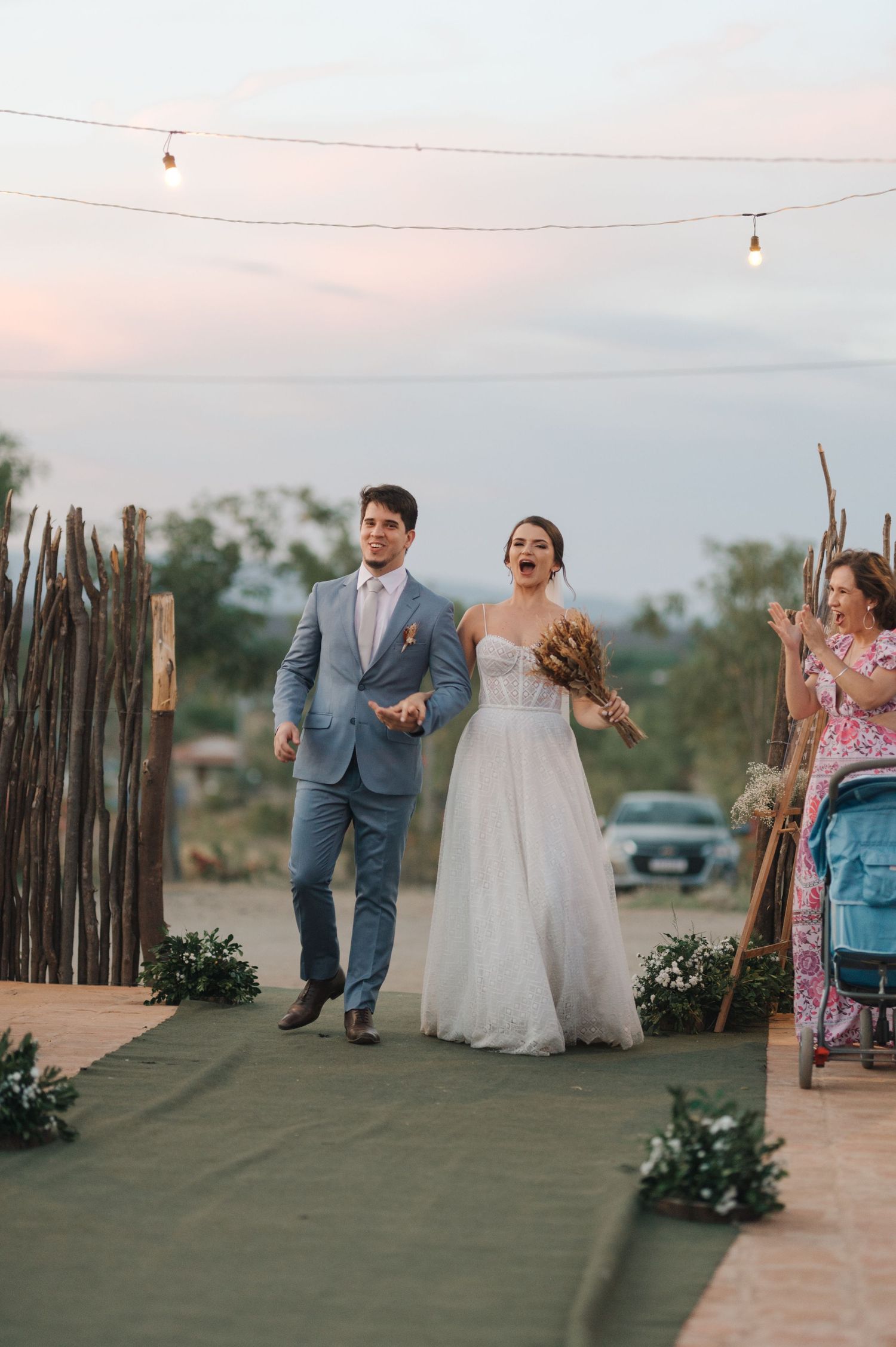Couple walking down an outdoor aisle at sunset with string lights and rustic decor.