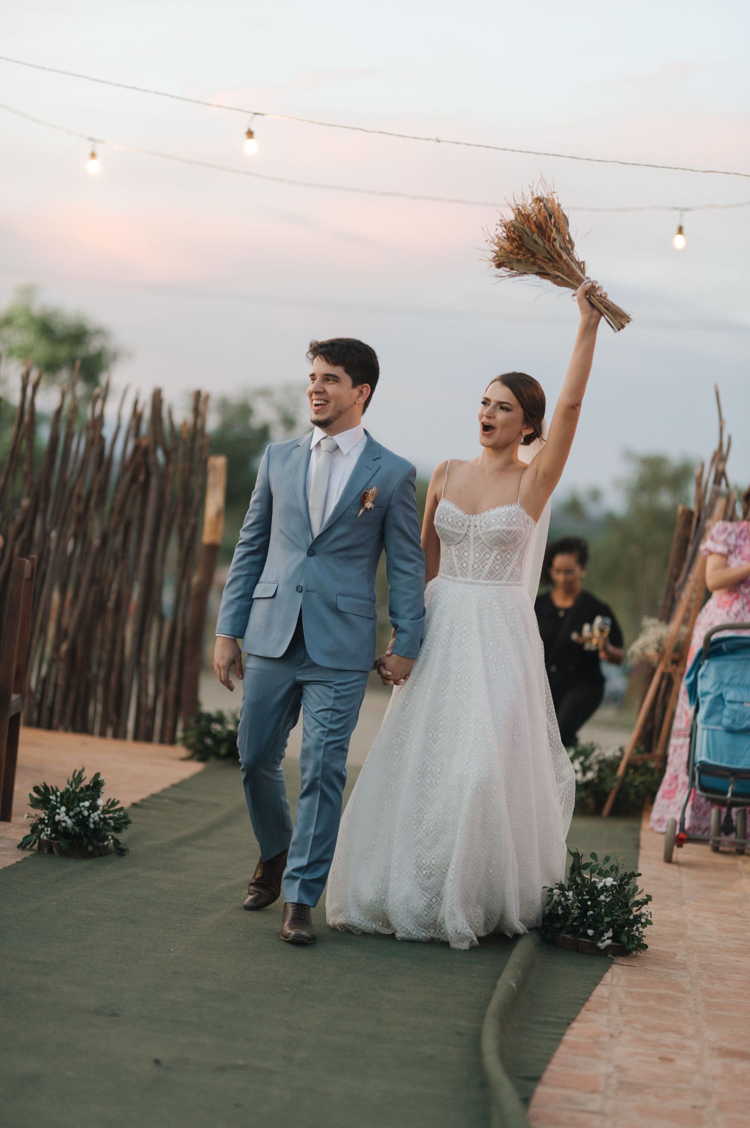 Couple celebrating during wedding ceremony with rustic outdoor backdrop and string lights overhead.