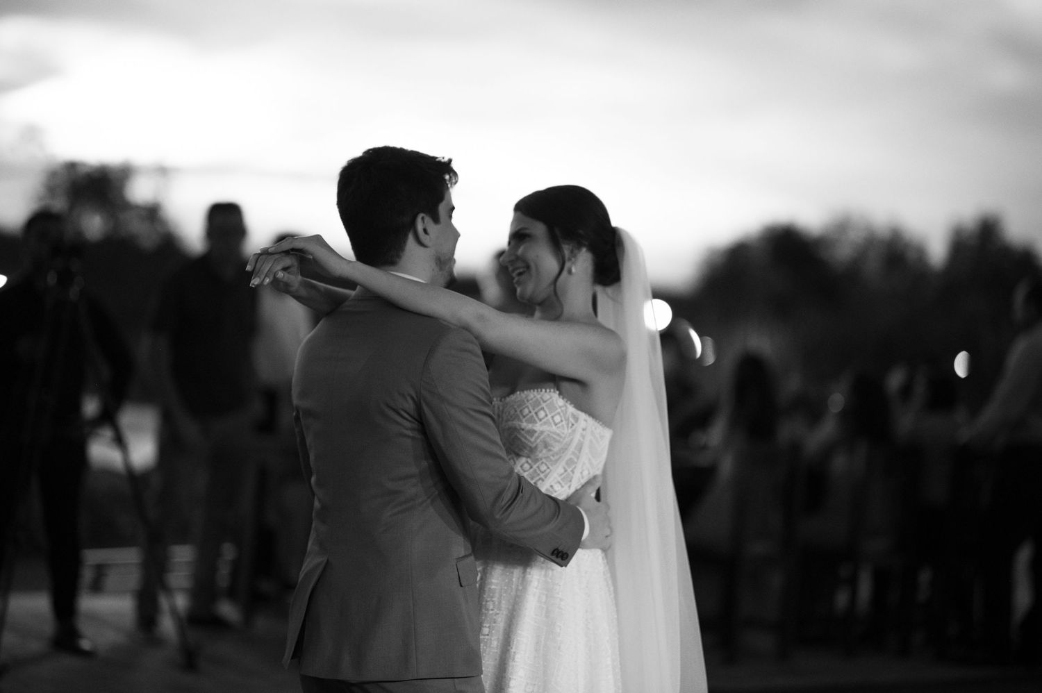 Black and white capture of couple dancing outdoors with wedding guests in background.