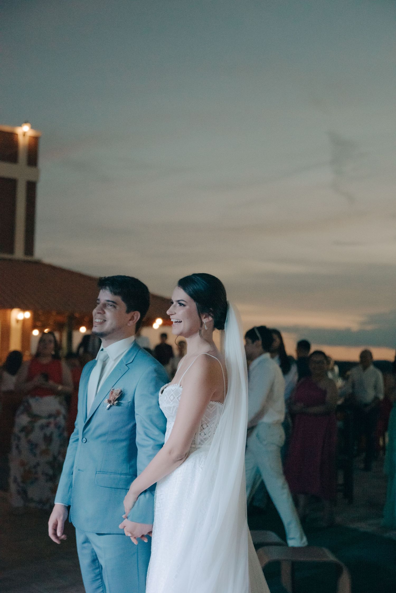 Couple in light blue suit and white dress stand together at sunset outdoor reception.