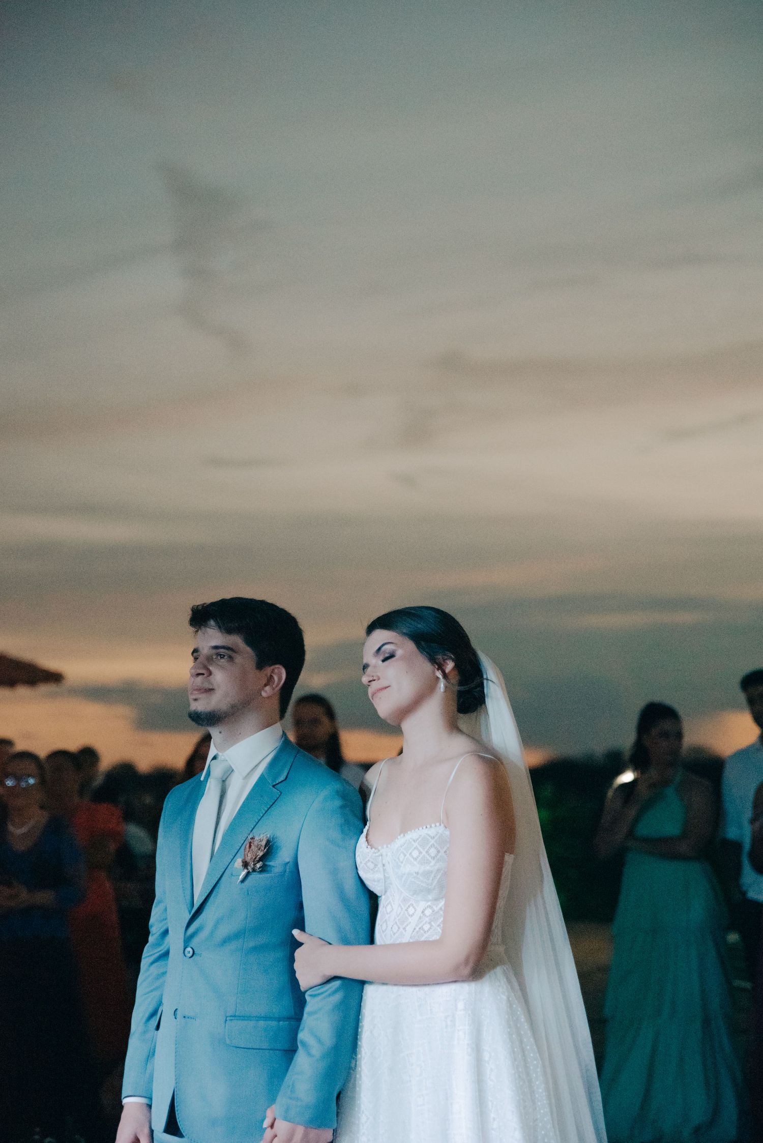 Couple in formal attire stands during sunset ceremony with guests in background.