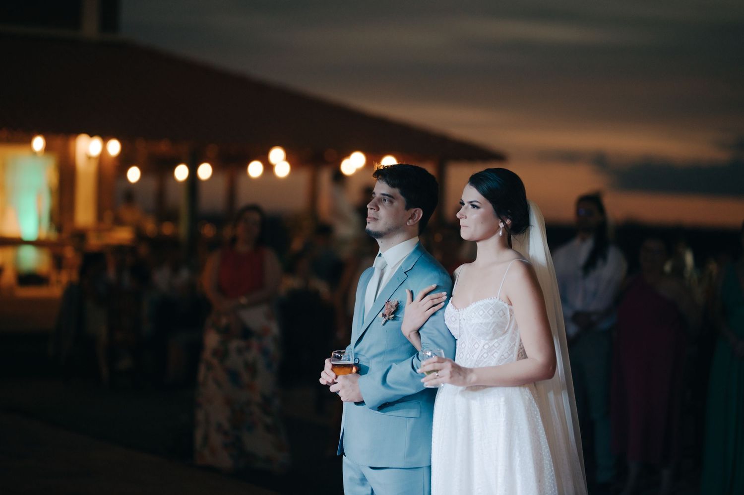 A couple stands together at an evening reception with glowing lights and guests in the background.